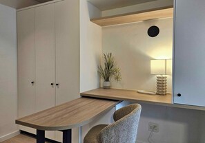 Una habitación con escritorio, silla, armario, planta, lámpara y una lámpara de pared.