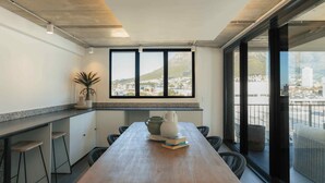 A modern kitchen with a wooden table, bar stools, and a view of a cityscape through large windows. - TEN on V - Fluent Serviced Apartments (Cape Town)