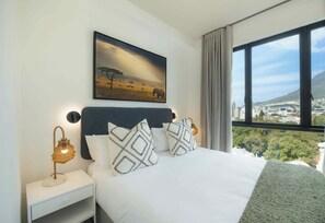 A hotel room with a bed, a nightstand, a wall-mounted lamp, a painting, and a window with a city view. - TEN on V - Fluent Serviced Apartments (Cape Town)