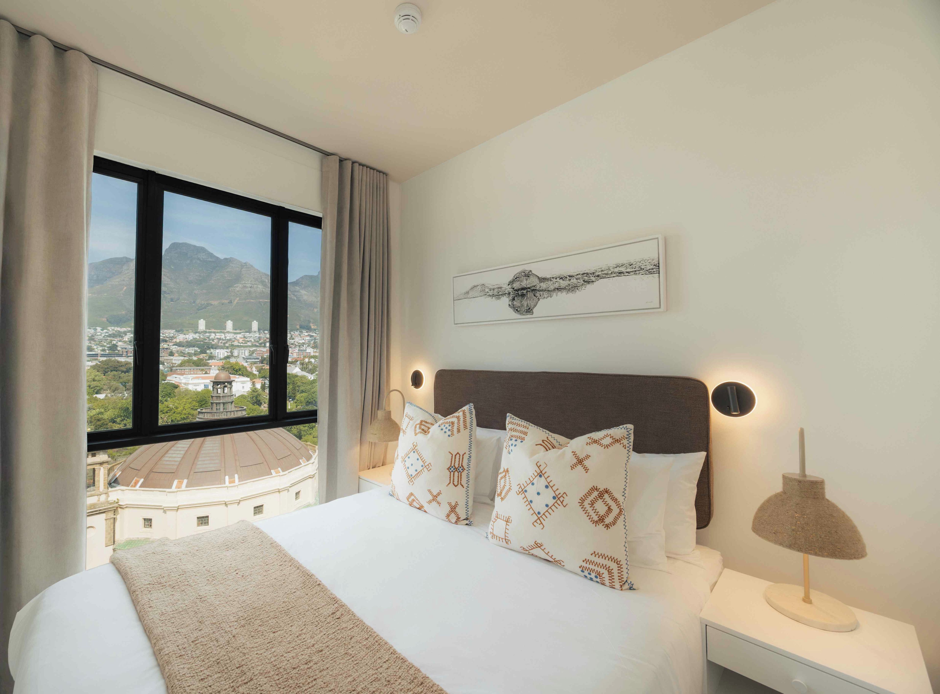 A hotel room with a large bed, a view of a cityscape, a mounted artwork, and a nightstand with a lamp.