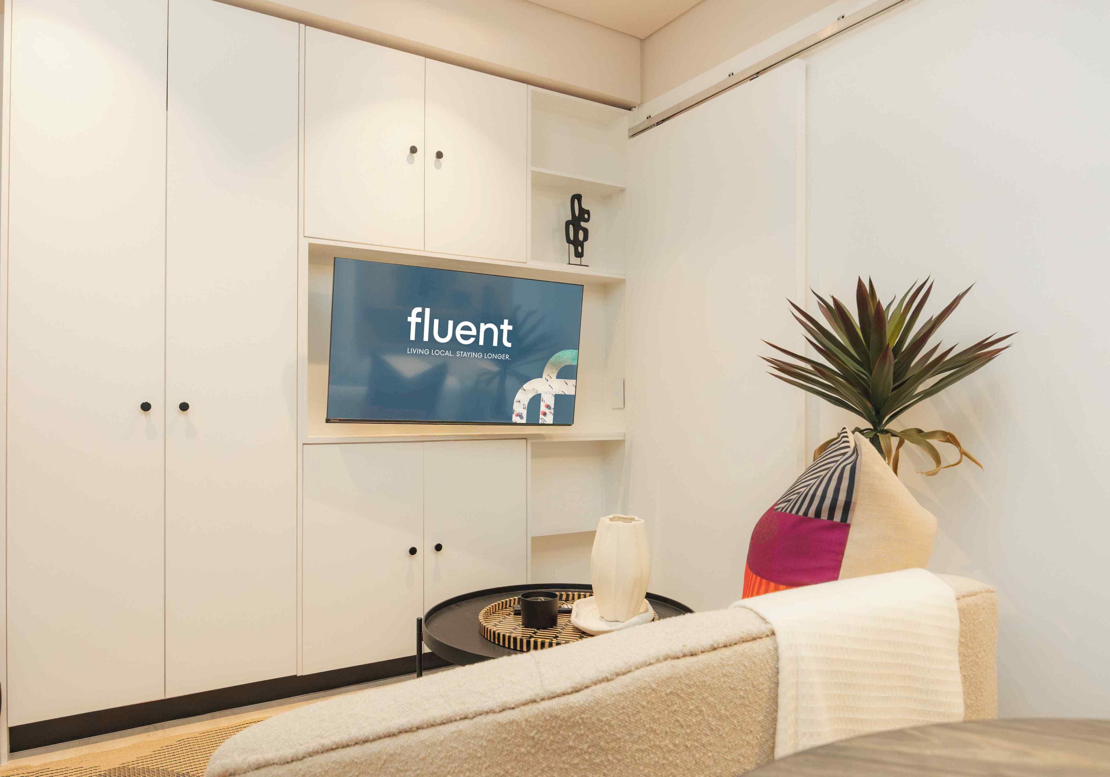 Una habitación con un sofá, una pequeña mesa con una vela, una planta en maceta y un televisor de pared que muestra la palabra 'fluent'.
