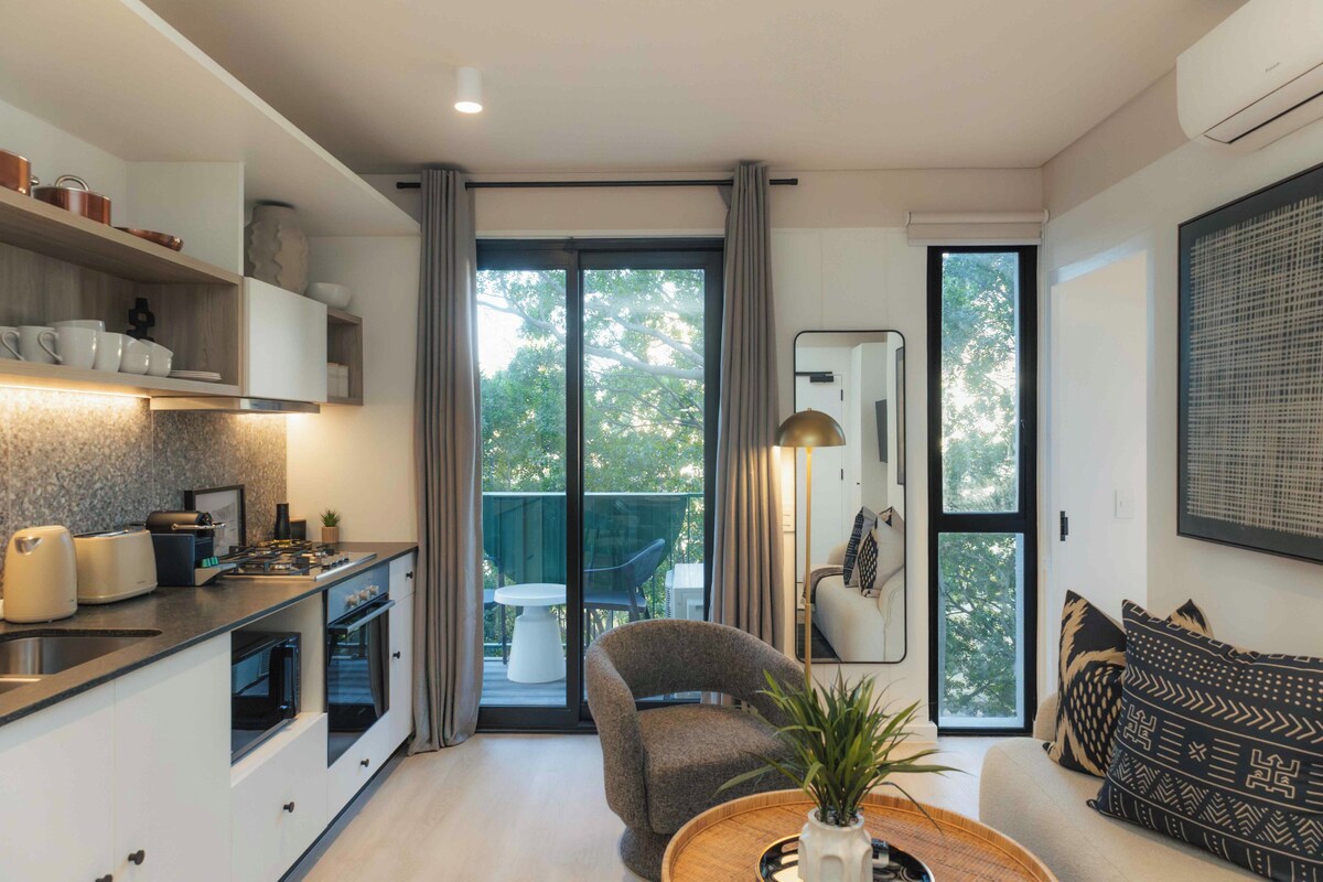 A modern kitchen with a dining area, a sofa, and a balcony with a view of trees.