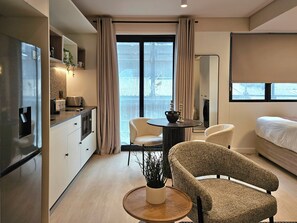 Quarto de hotel moderno com cozinha compacta, área de refeições com mesa redonda e cadeiras, uma cama grande e vista através de uma porta de vidro deslizante.