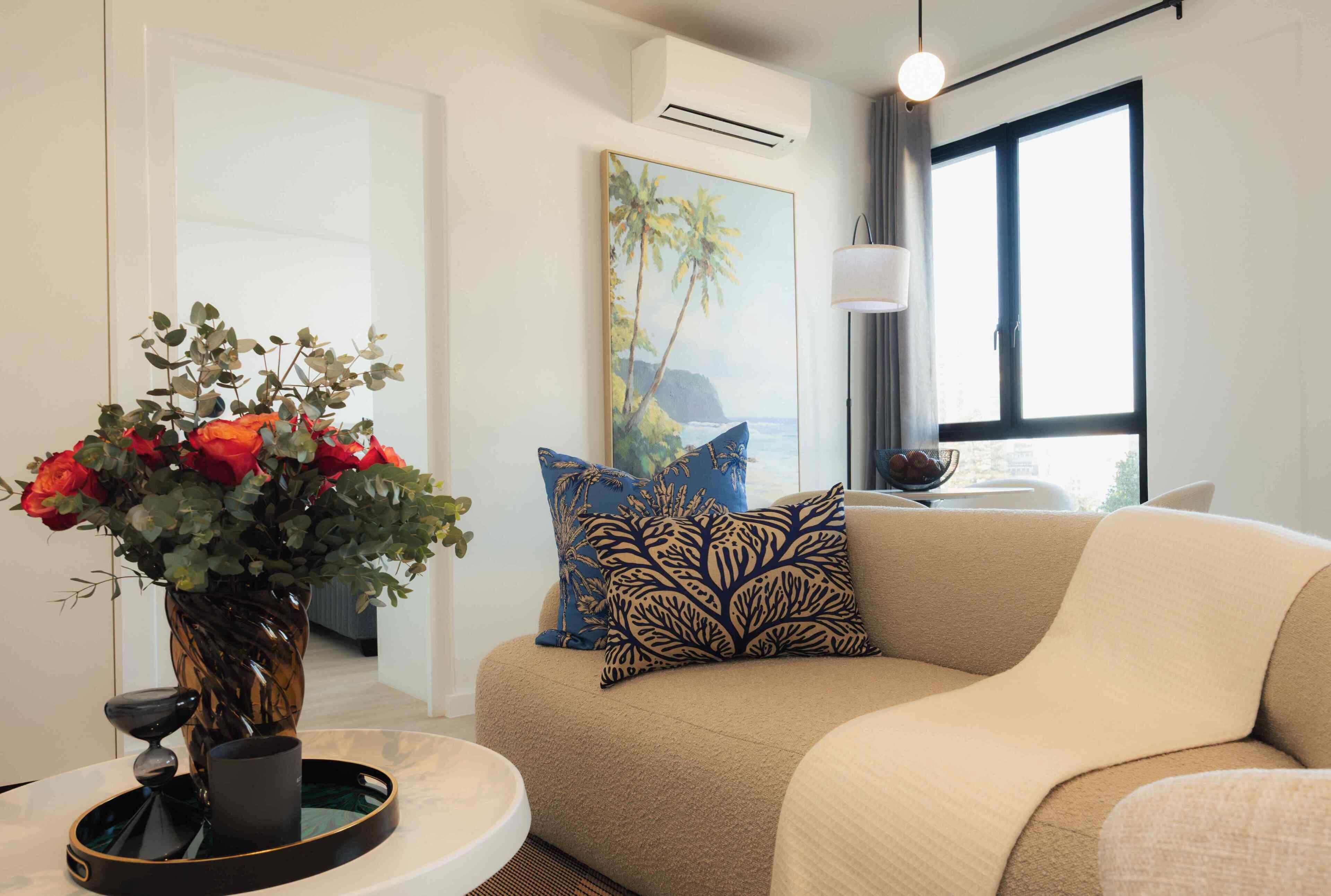 Un salon moderne avec un canapé beige, un vase de fleurs rouges et un tableau représentant un paysage tropical.