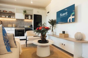 A modern living room with a sofa, a coffee table with a vase of flowers, a kitchen in the background, and a wall-mounted TV displaying the word 'fluent'. - TEN on V - Fluent Serviced Apartments (Cape Town)
