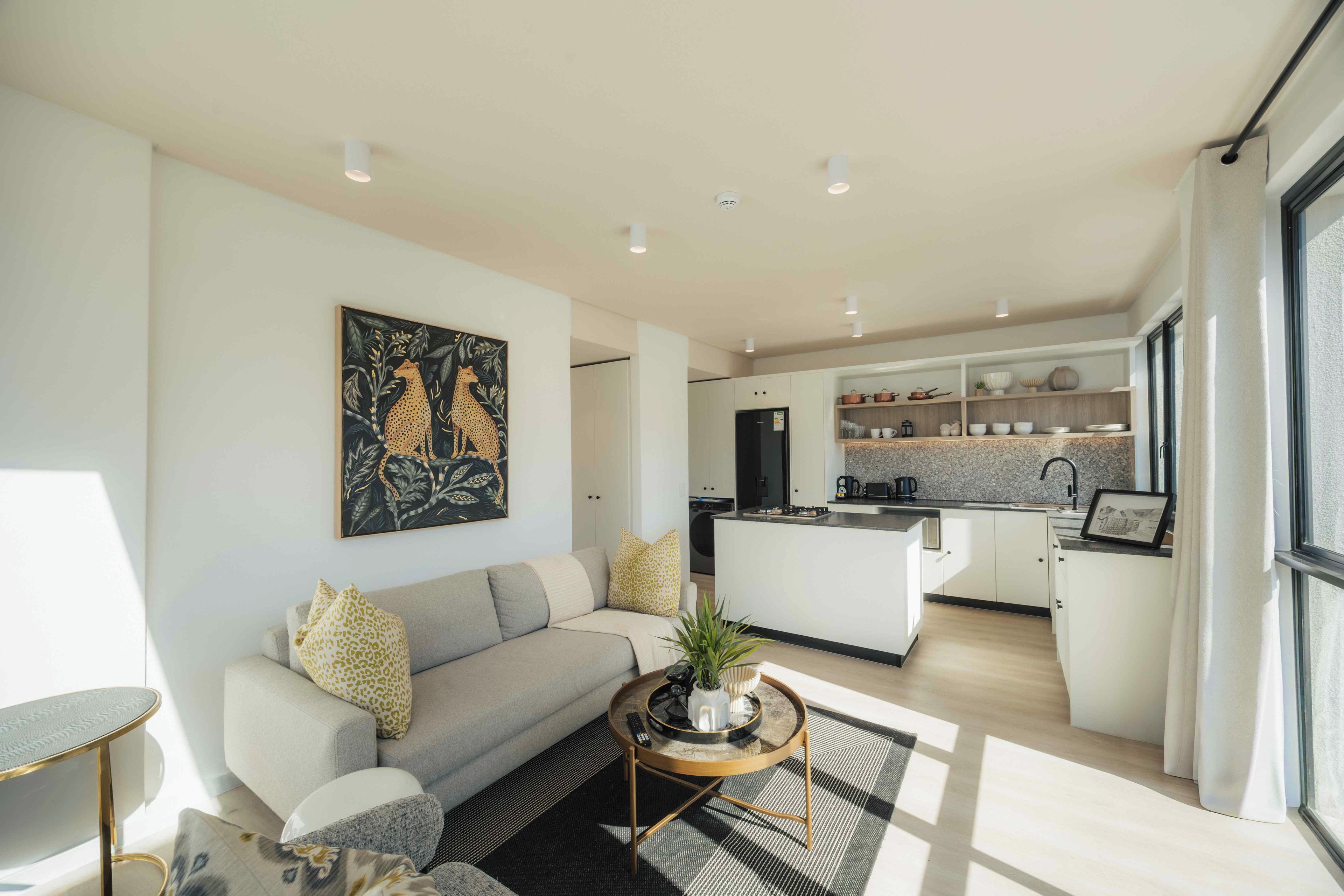 A modern living room with a sofa, a coffee table, and a painting. There is a kitchen area with a sink and cabinets in the background.