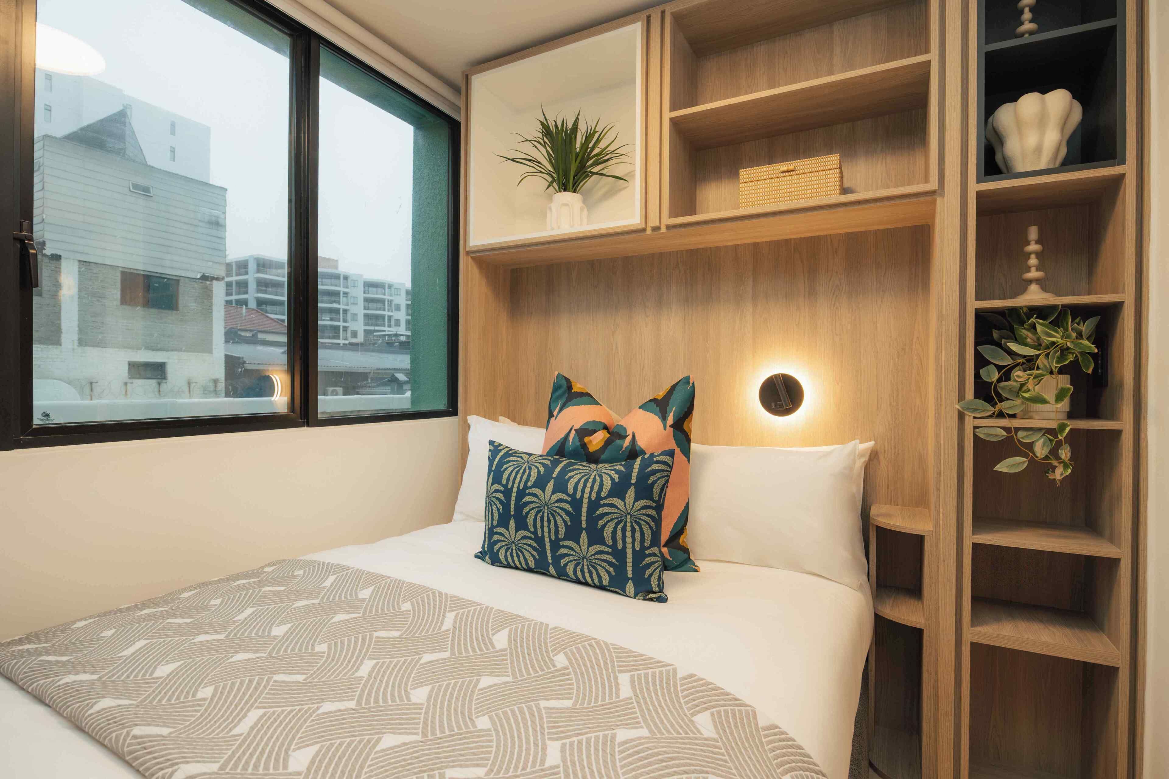 A hotel room with a bed, a bedside table, a nightstand, a shelf with decorative items, and a window with a view of buildings.