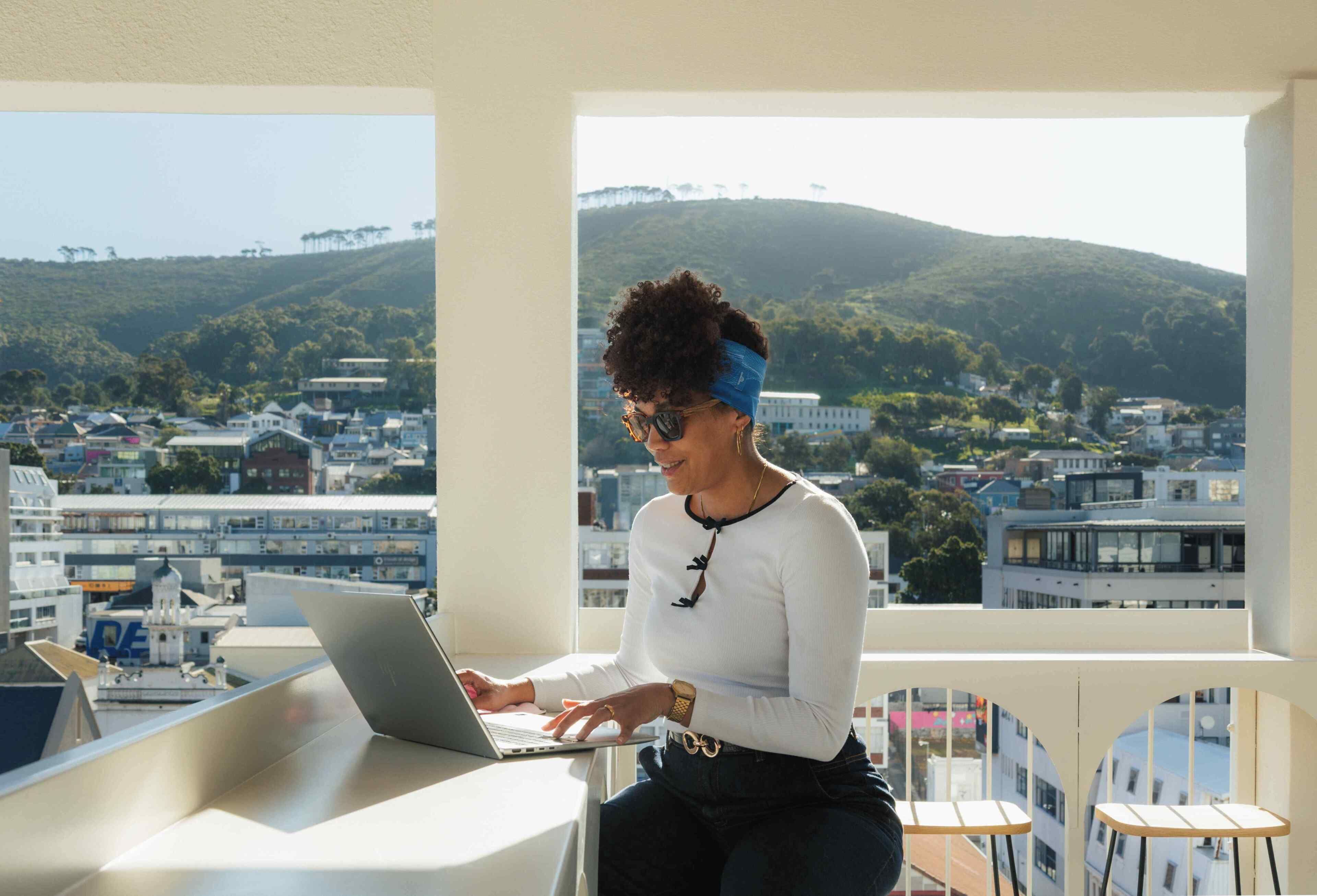 A person using a laptop on a balcony with a cityscape and hills in the background.