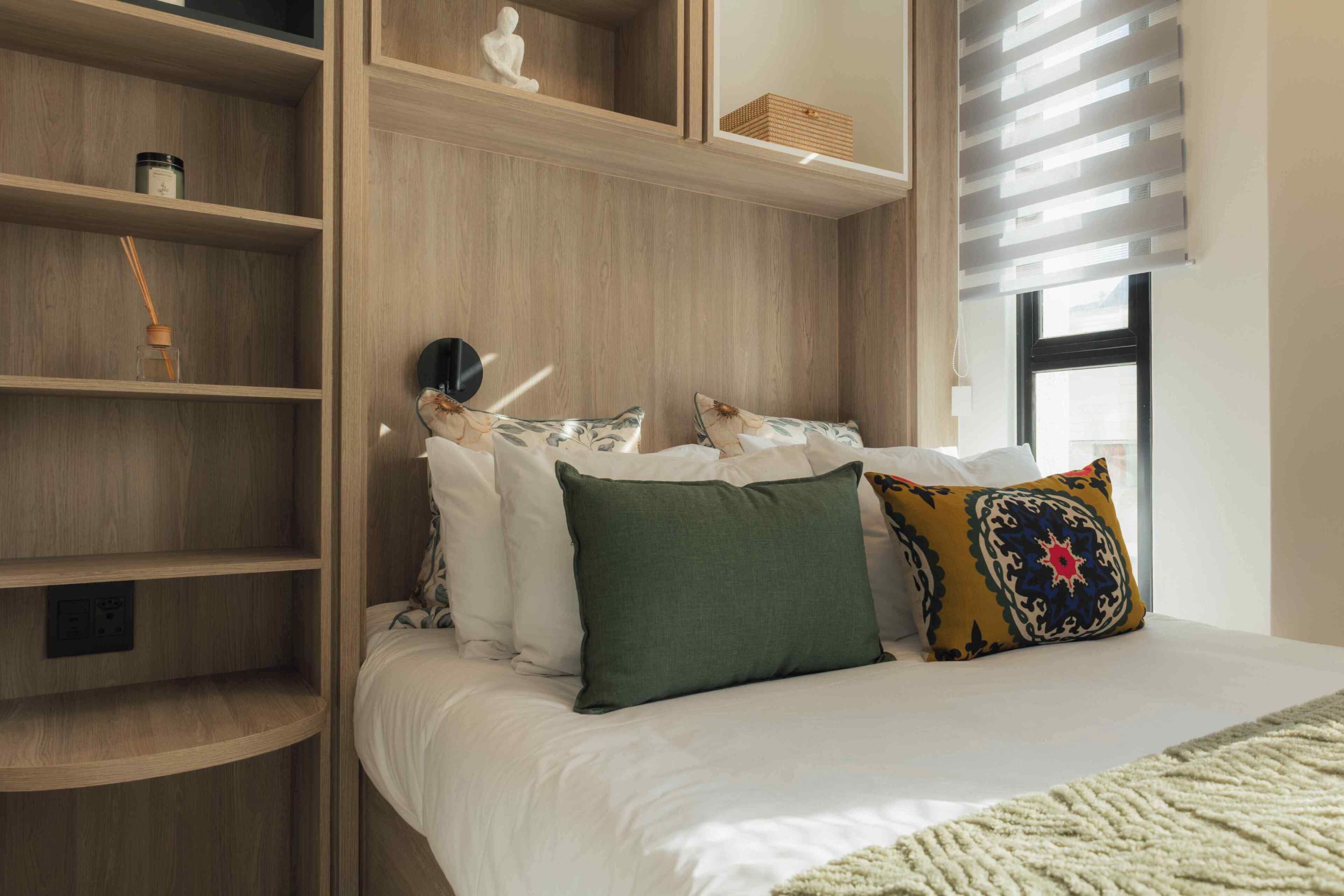 A bedroom with a bed, a nightstand, a shelf with a decorative item, and a window with blinds.