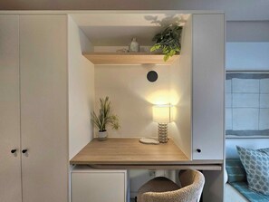 Habitación pequeña con escritorio, lámpara, planta, estante con adornos y una cama para mascotas.