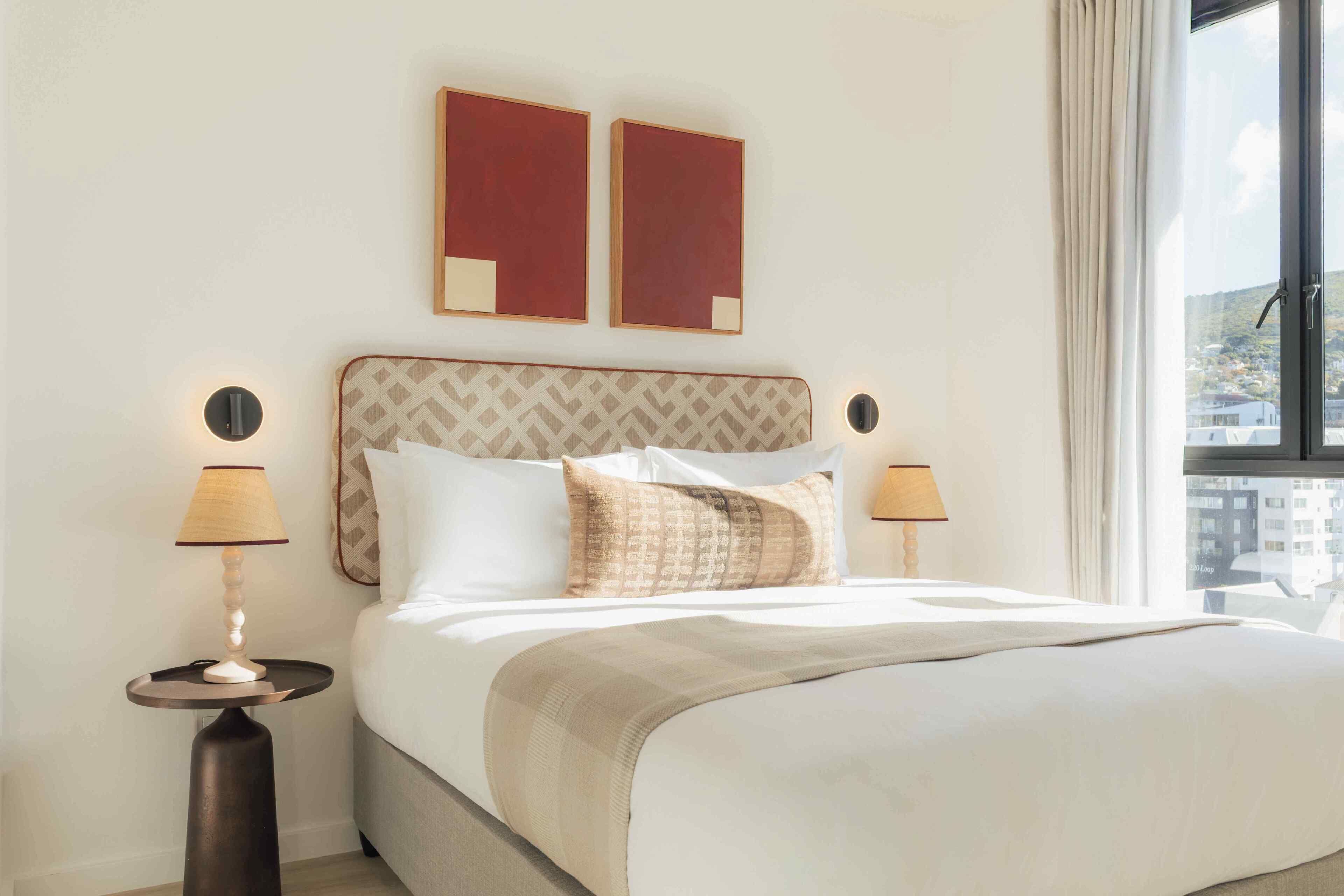 A hotel room with a bed, two bedside tables, two lamps, a window with curtains, and two wall-mounted artworks.