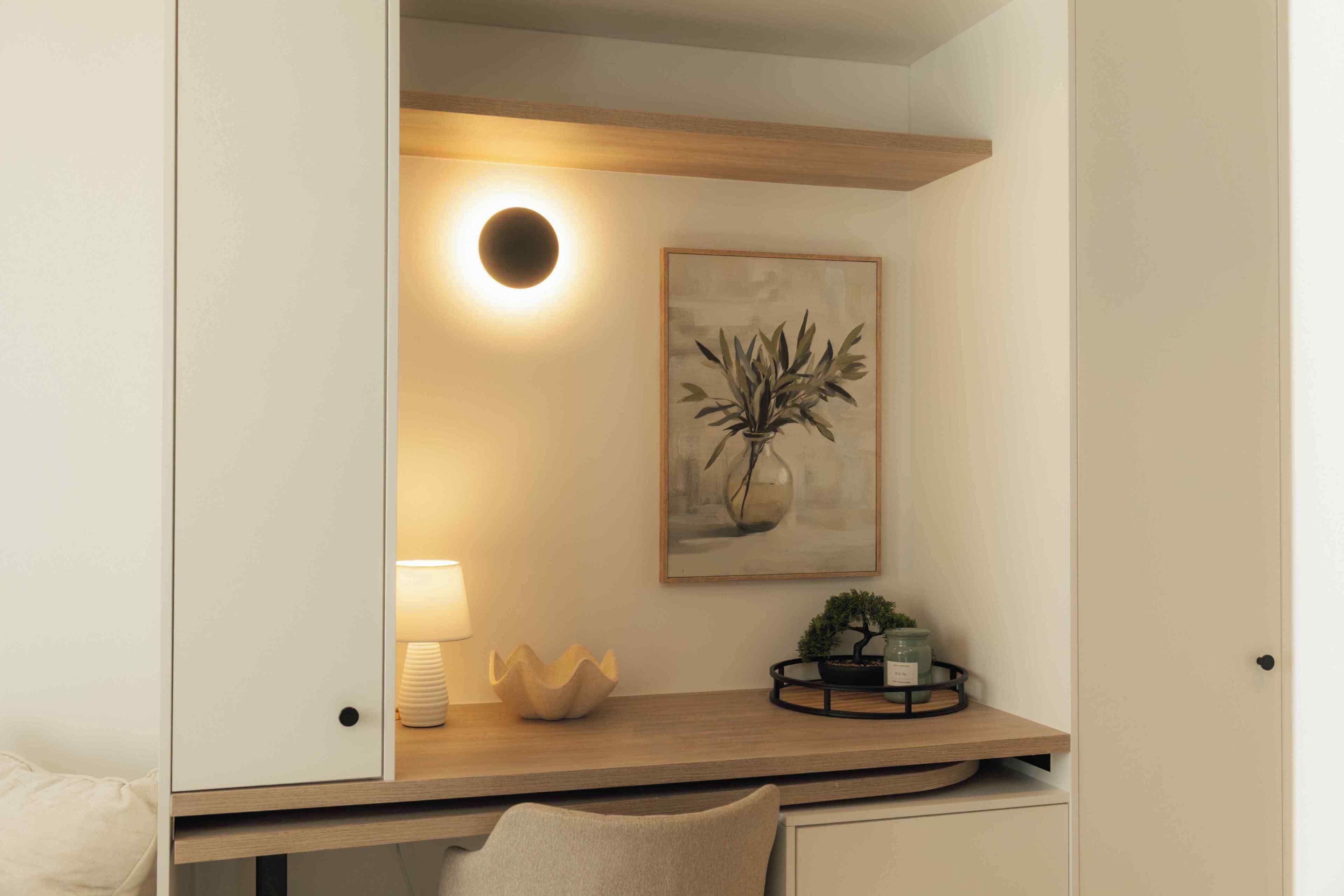 A room with a desk, a lamp, a framed artwork, and a small potted plant.