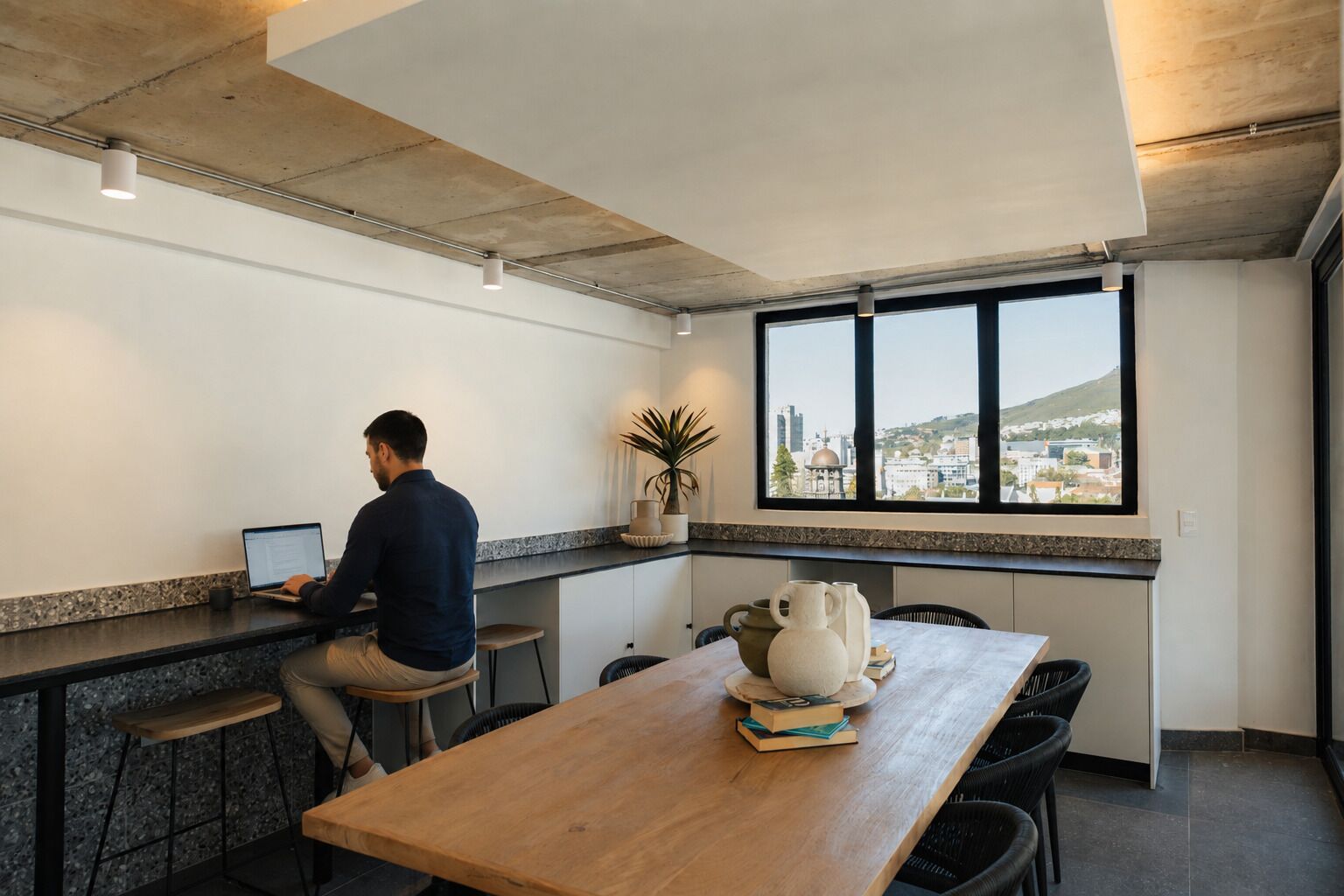A person working at a desk with a laptop in a modern kitchen area with a view of a cityscape.