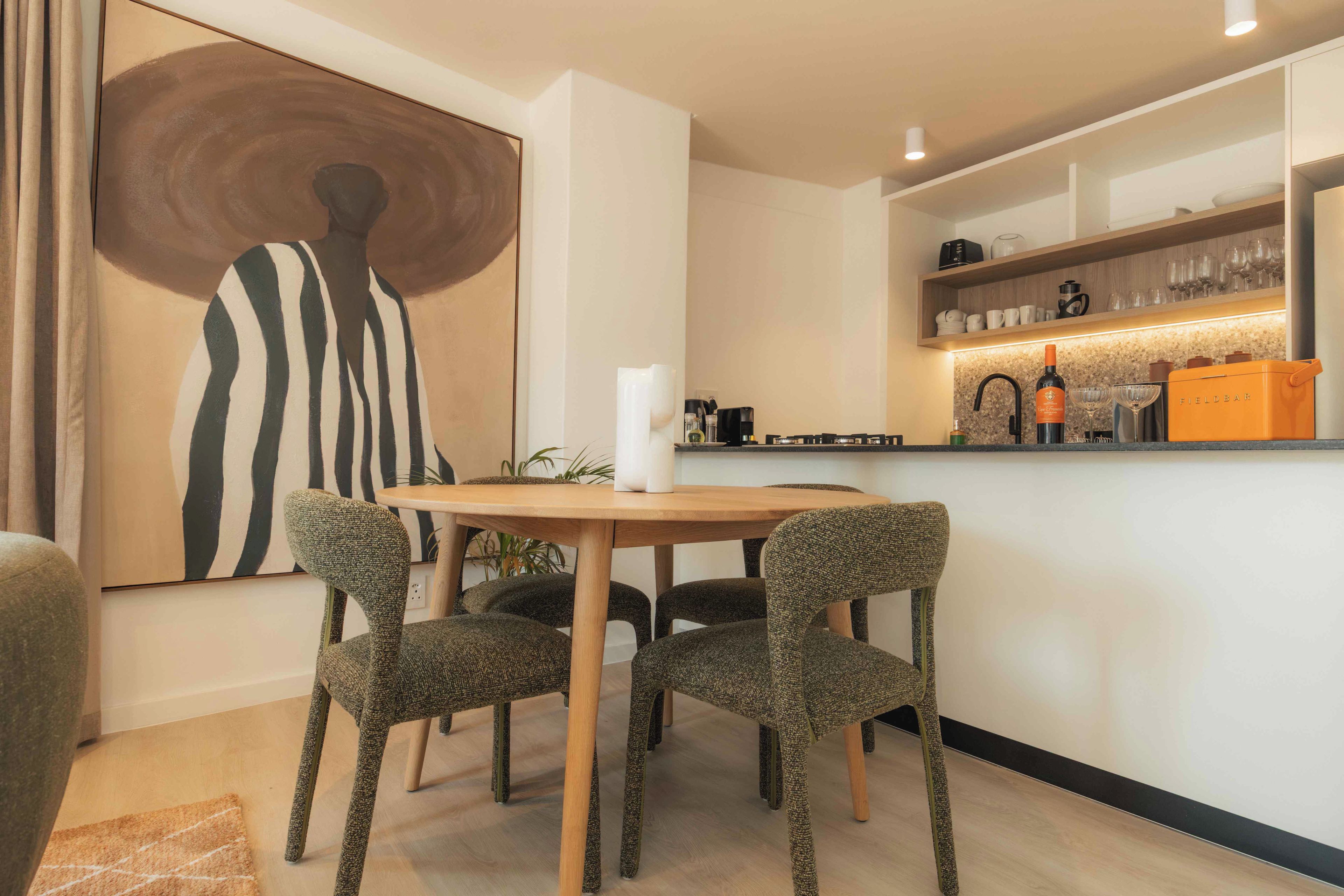 Zona pranzo moderna con tavolo rotondo, quattro sedie, un grande quadro astratto e un bancone cucina con diversi oggetti.