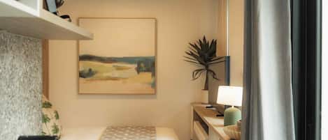 A hotel room with a bed, a desk, a lamp, a plant, and a painting on the wall.