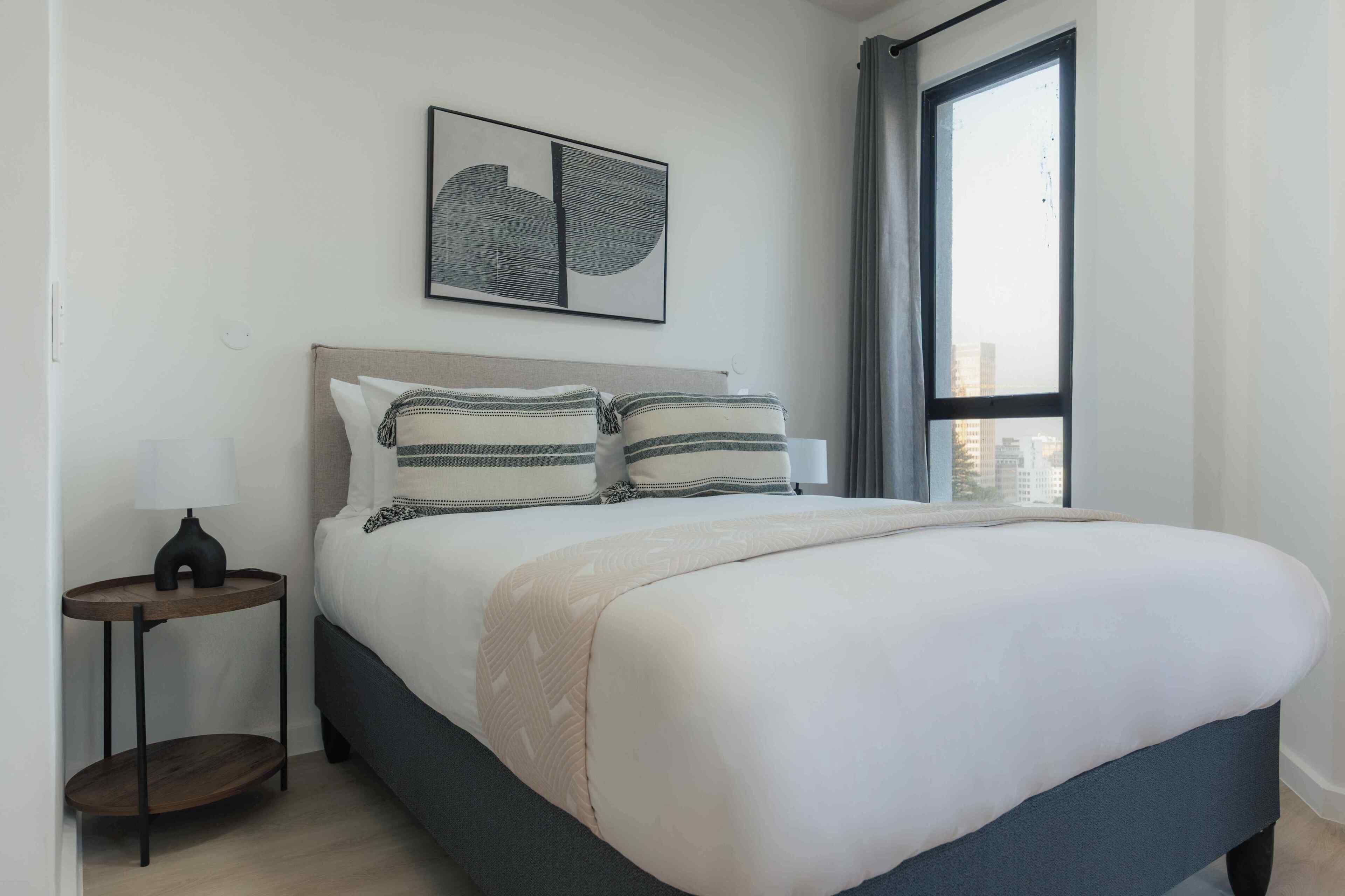 A bedroom with a bed, a nightstand, a lamp, a window with curtains, and a wall-mounted artwork.