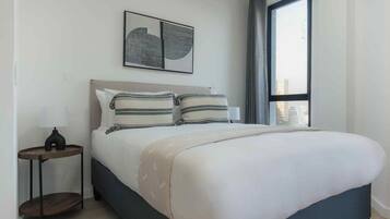 A bedroom with a bed, a nightstand, a lamp, a window with curtains, and a wall-mounted artwork.