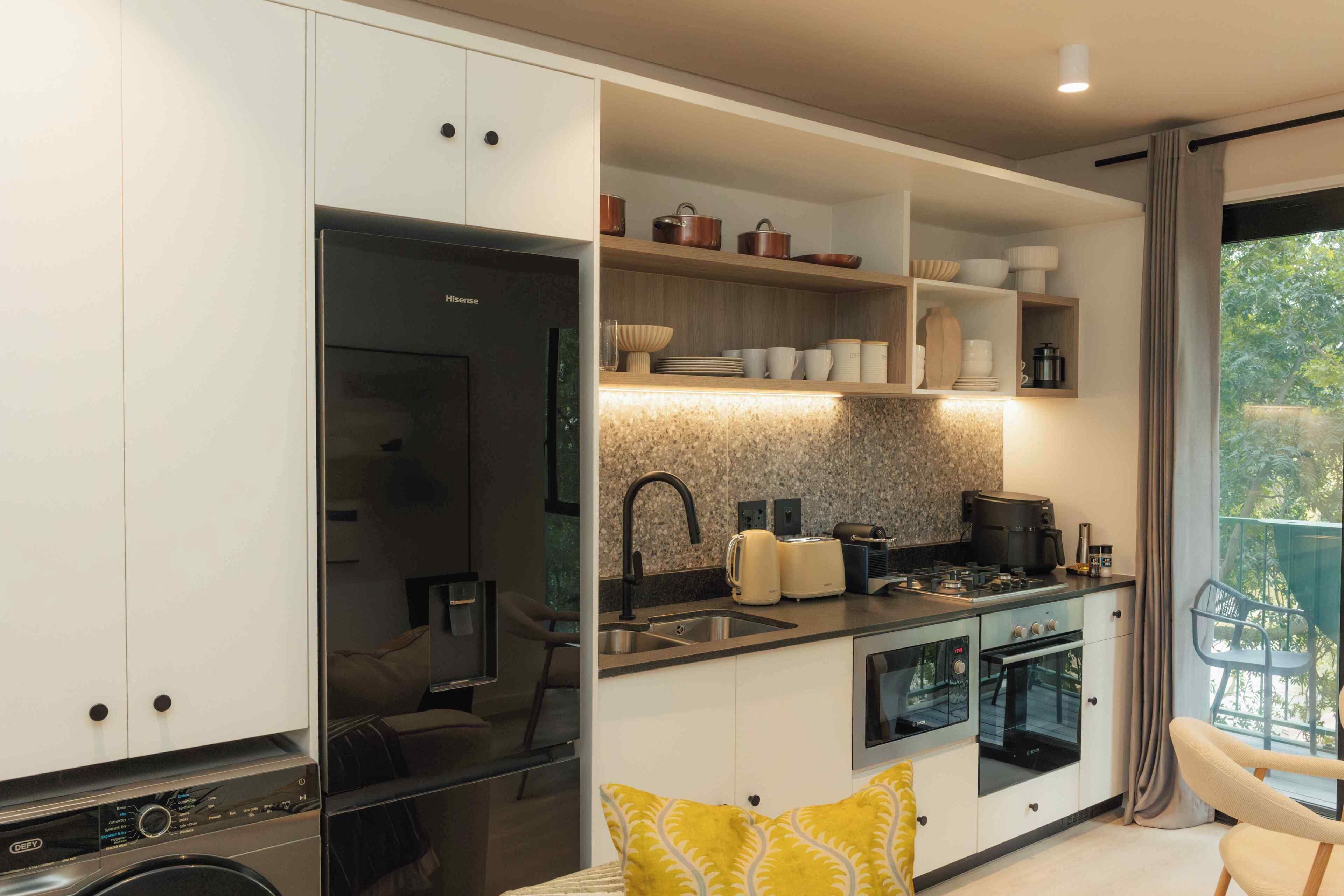 Cocina compacta con lavadora incorporada, zona de comedor con un cojín amarillo y vistas a la vegetación por la ventana.