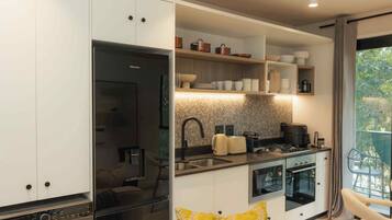 A compact kitchen with a built-in washing machine, a dining area with a yellow cushion, and a view of greenery through the window.