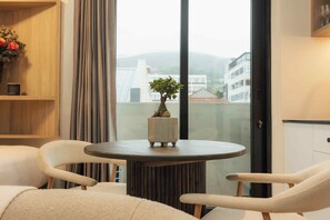 Una habitación con una mesa redonda, dos sillas, una planta en maceta y vistas de edificios a través de una ventana.