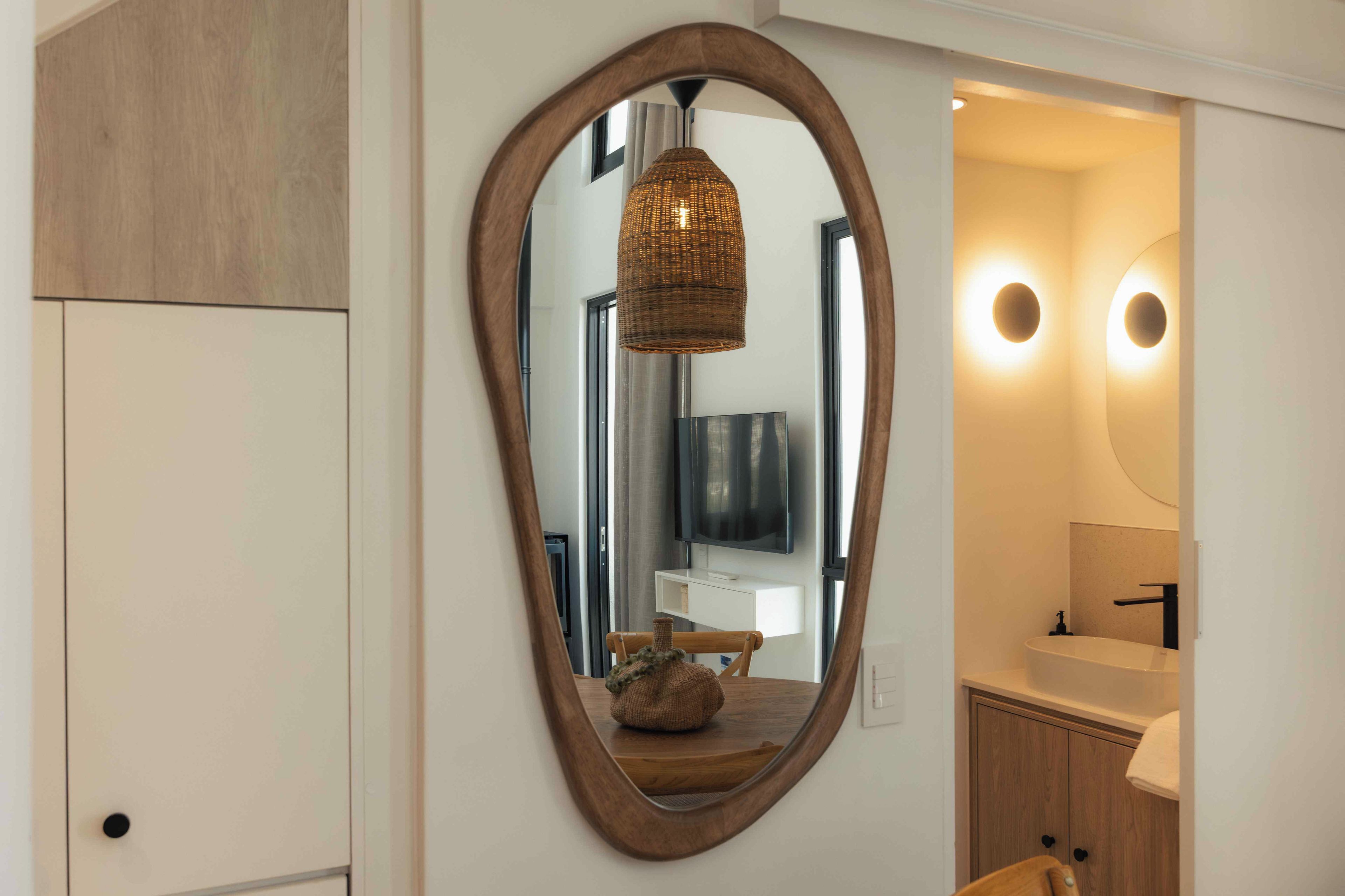 Et spejl med oval ramme, en fletlampe, et fjernsyn og et badeværelse med vask og skab.