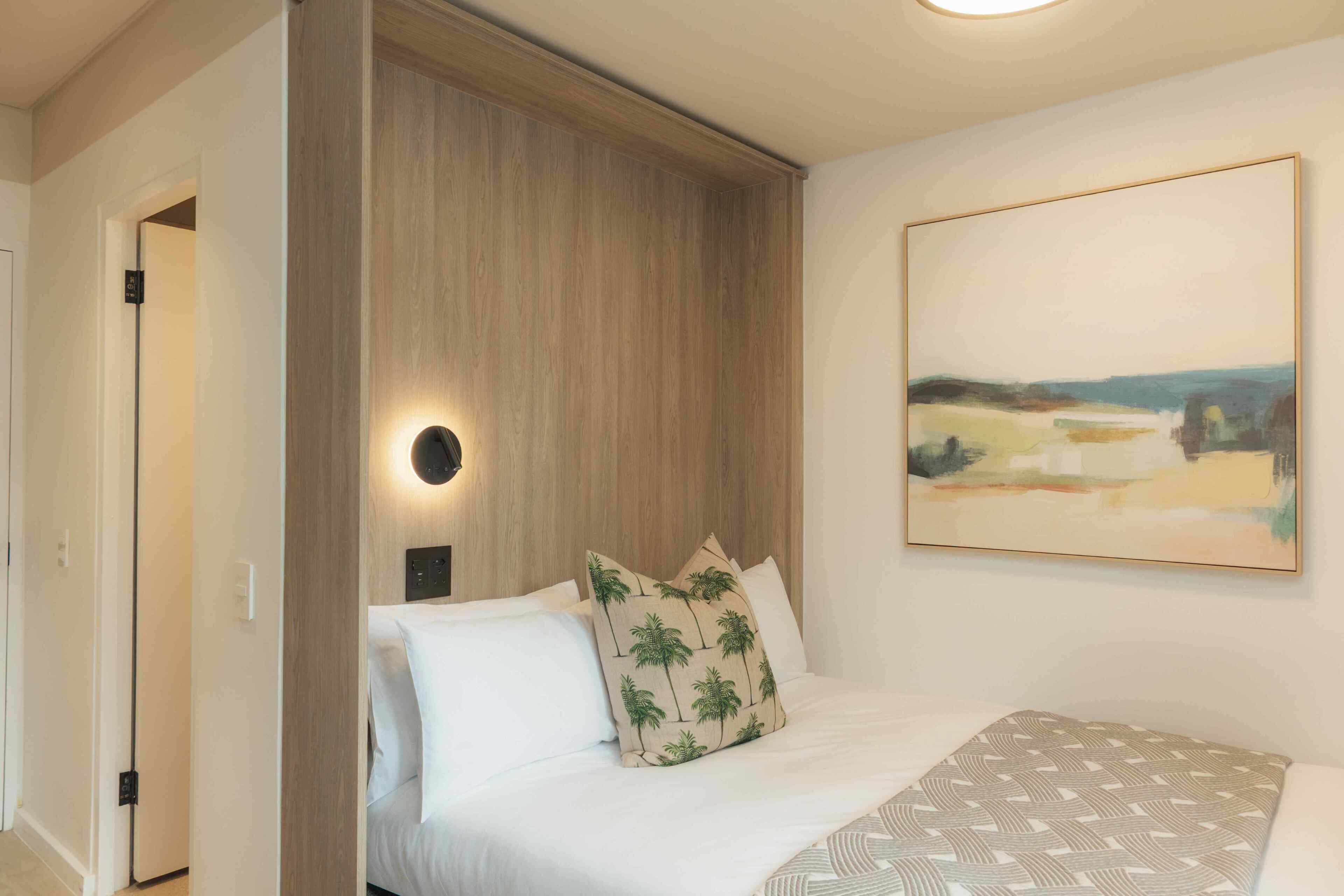 Une chambre à coucher avec un lit, une table de chevet, une lampe fixée au mur et un tableau.