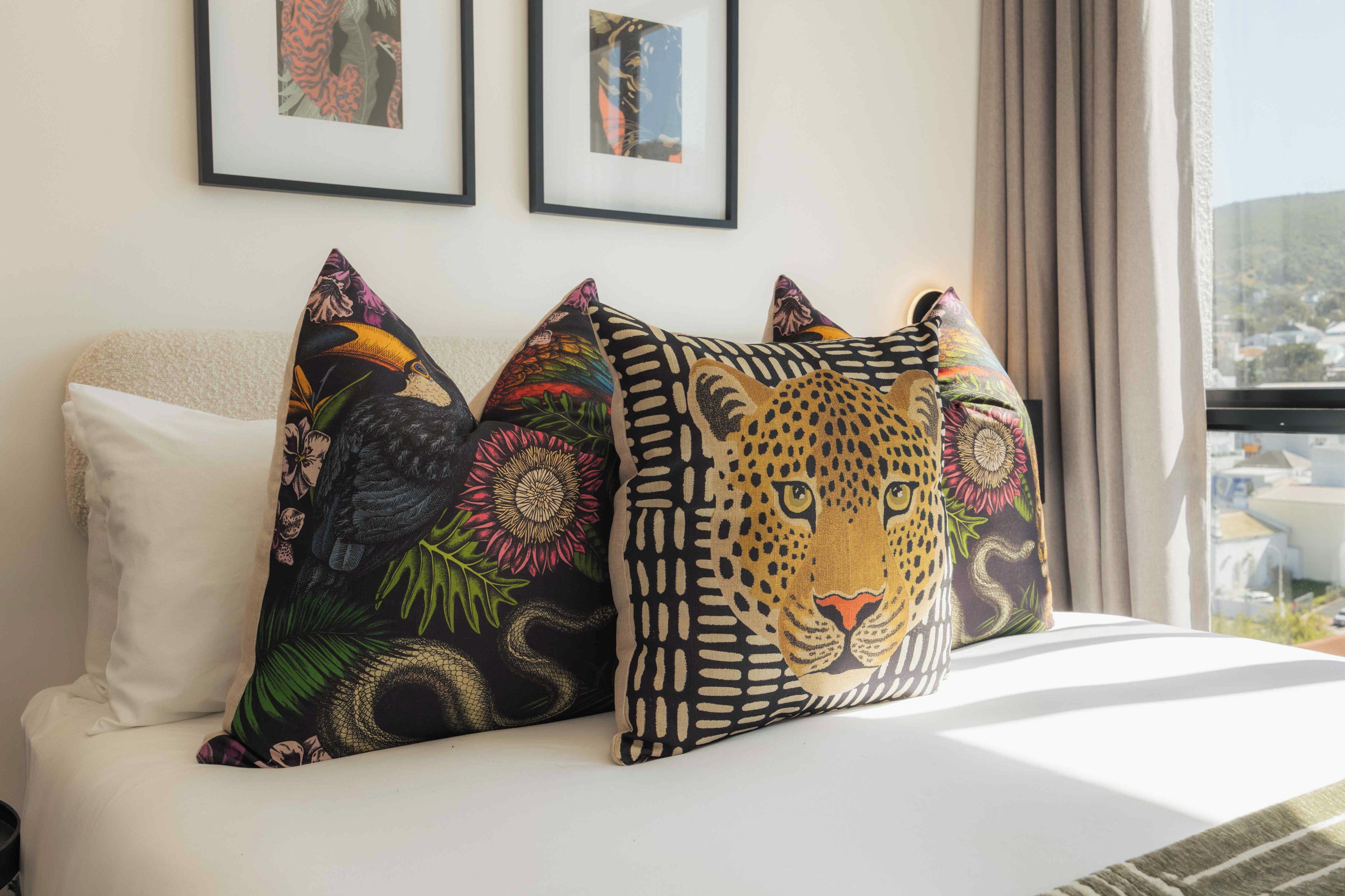 Ein Bett mit mehreren Kissen, darunter ein auffälliges Kissen mit Leopardenmuster, in einem Zimmer mit gerahmten Bildern an der Wand und Blick durch ein Fenster.
