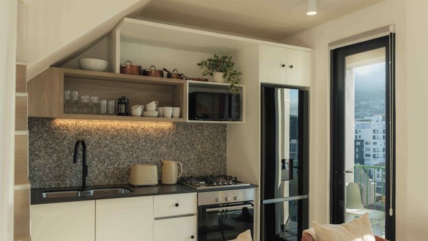 A compact kitchen with a built-in microwave, stove, and oven. There is a sink with a modern faucet, and a countertop with a kettle and toaster. A plant is on the shelf above the microwave. - TEN on V - Fluent Serviced Apartments (Cape Town)