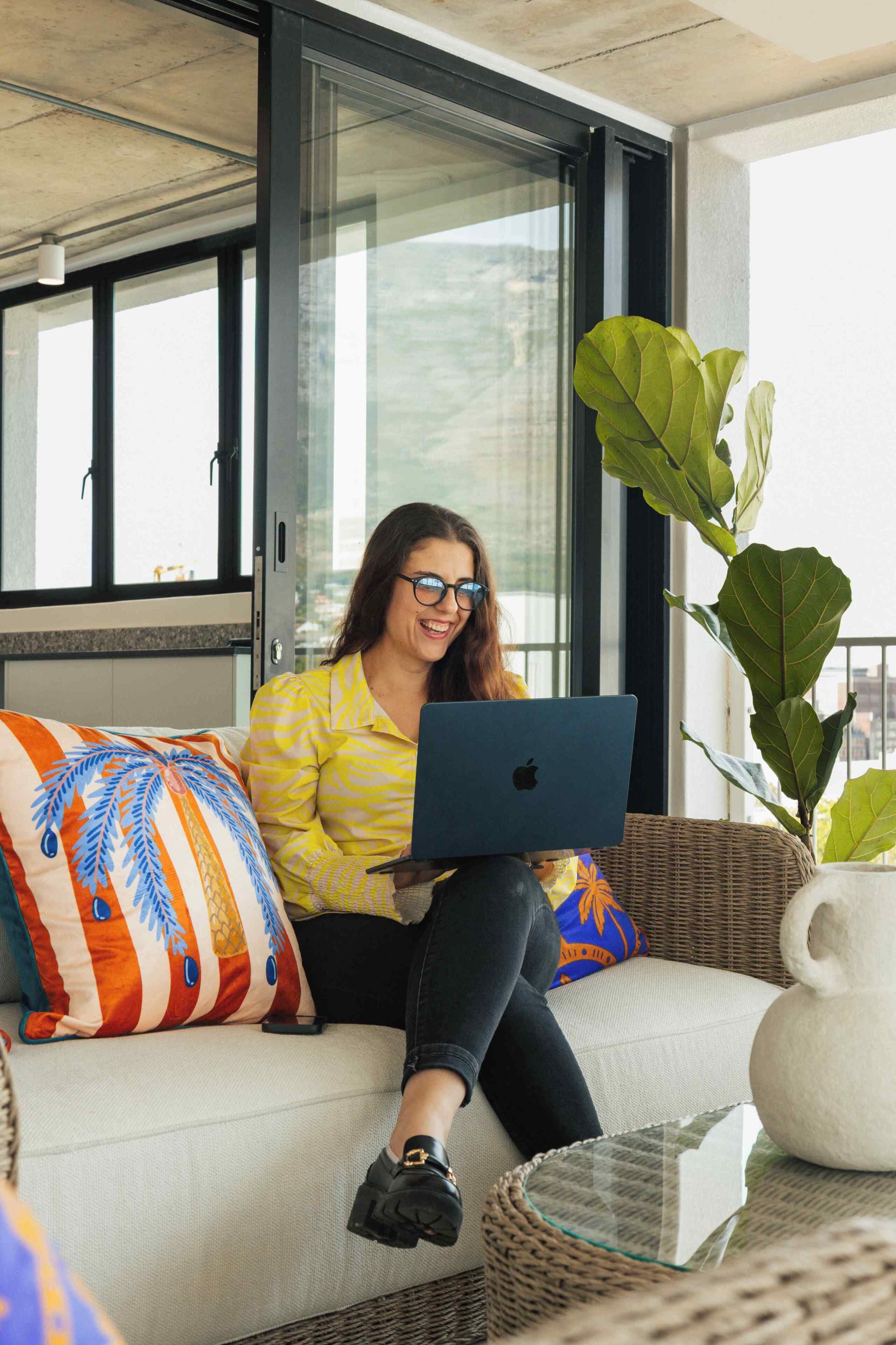 A person sitting on a sofa with a laptop, a large plant, and a glass table.