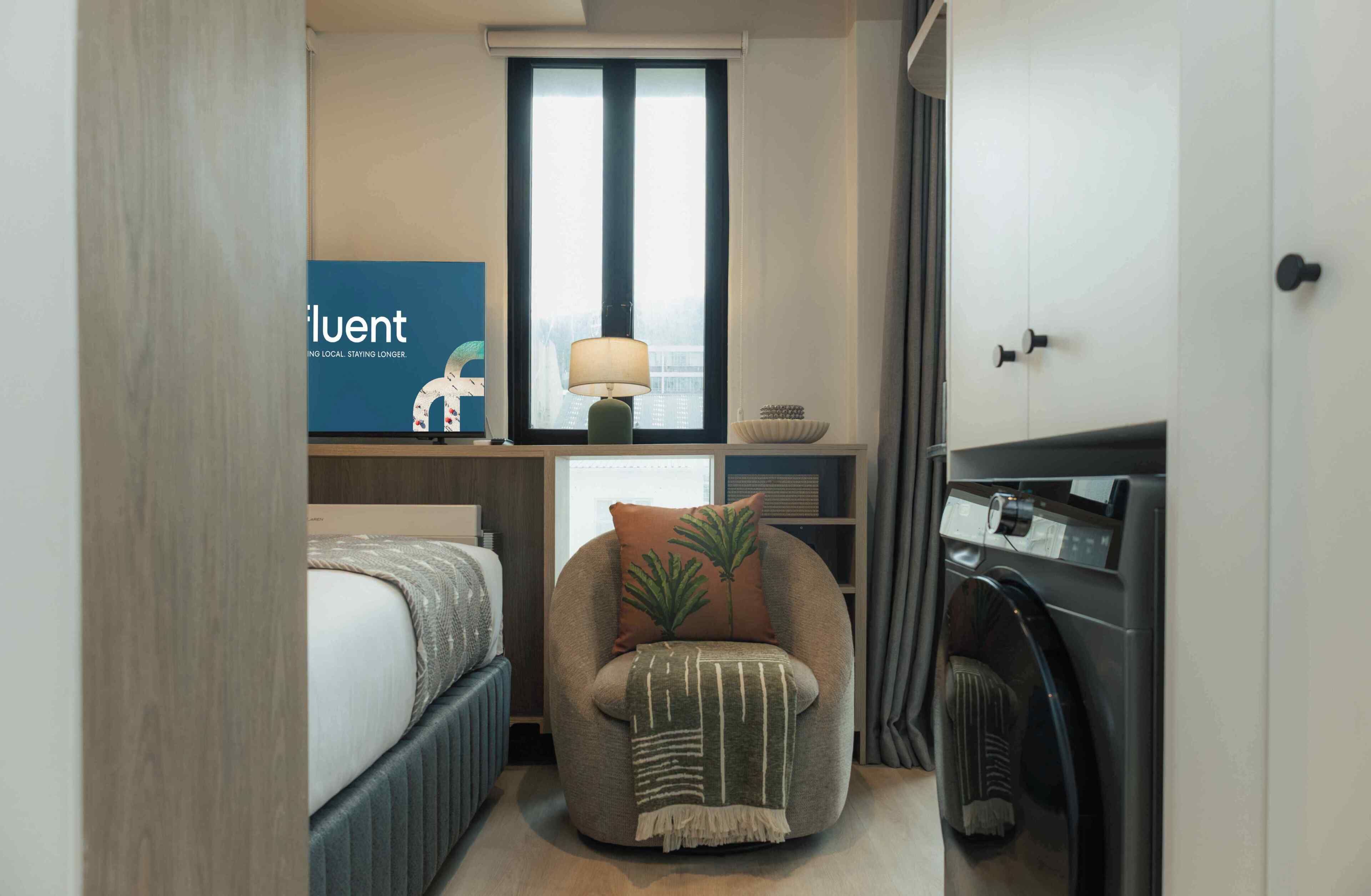 A compact hotel room with a bed, a chair with a blanket, a desk with a lamp, a window with blinds, and a washing machine.