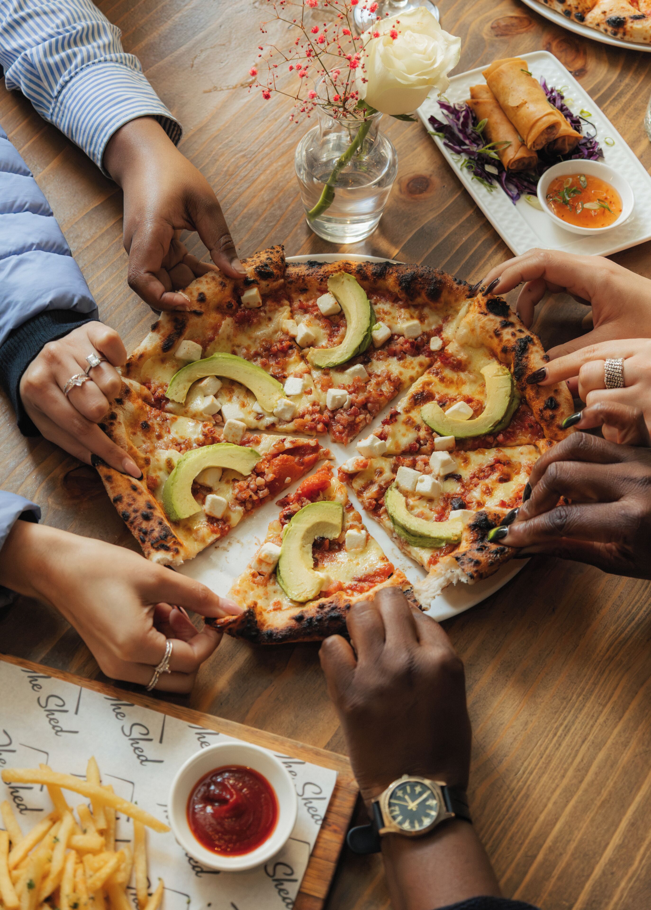 Een groep mensen rondom een pizza met avocado, omringd door verschillende gerechten en drankjes.