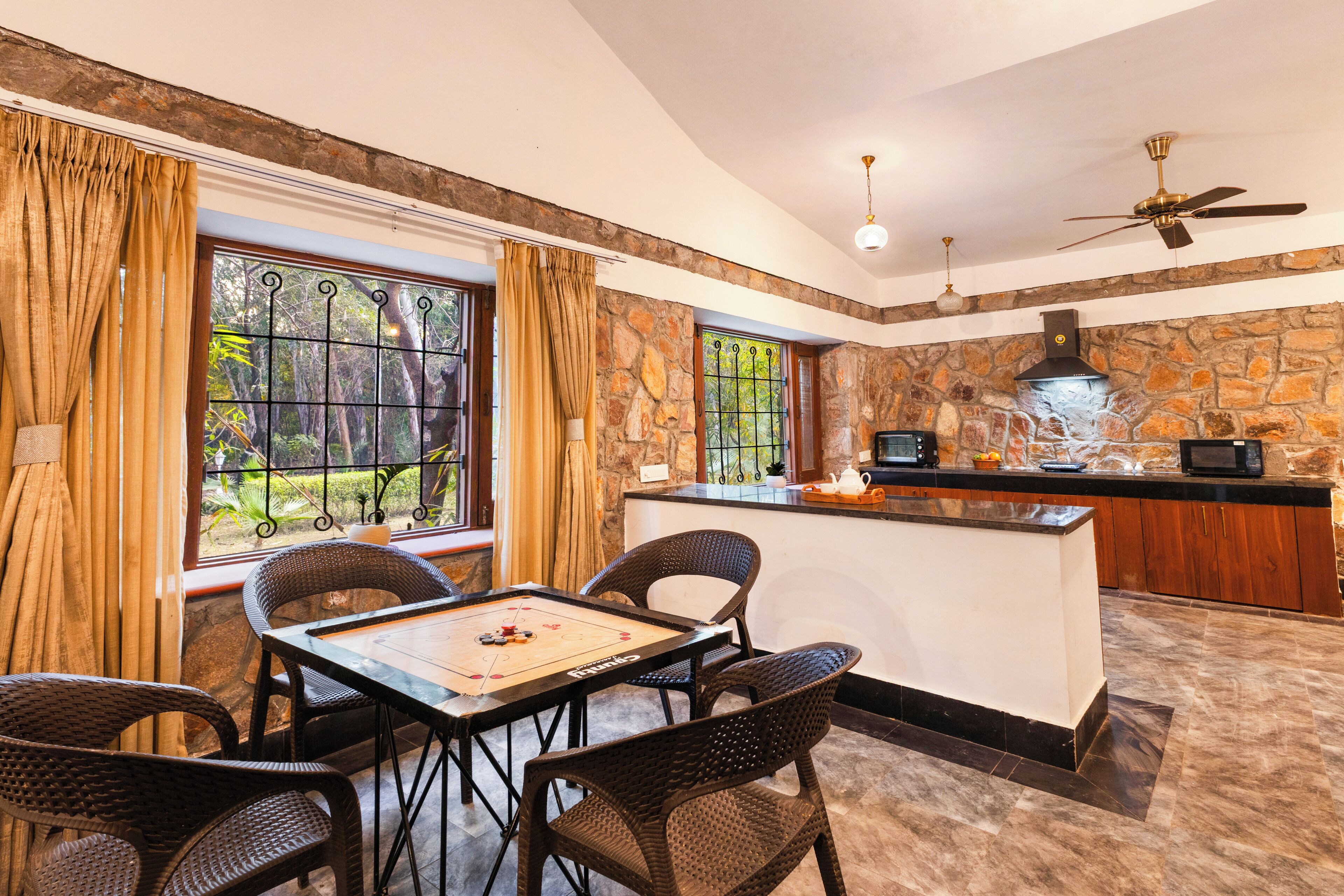 A dining area with a table and chairs, a kitchen with a stone wall, and a view of greenery through the windows.