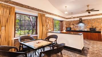 A dining area with a table and chairs, a kitchen with a stone wall, and a view of greenery through the windows.
