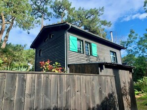 A two-story wooden house with a dark exterior and green shutters, surrounded by a wooden fence and trees. - 1st line to the Mimbeau lagoon in Cap Ferret A0340 (Lège-Cap-Ferret)