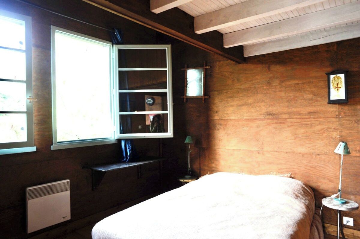 A room with a bed, a window with a view, a shelf with items, and a wall-mounted picture.