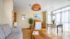 A living room with a sofa, a wooden coffee table, a dining table with chairs, a wicker chair, a potted plant, and a hanging light fixture. - Isla de Cuba 2K Apartment – Colombres (Colombres)
