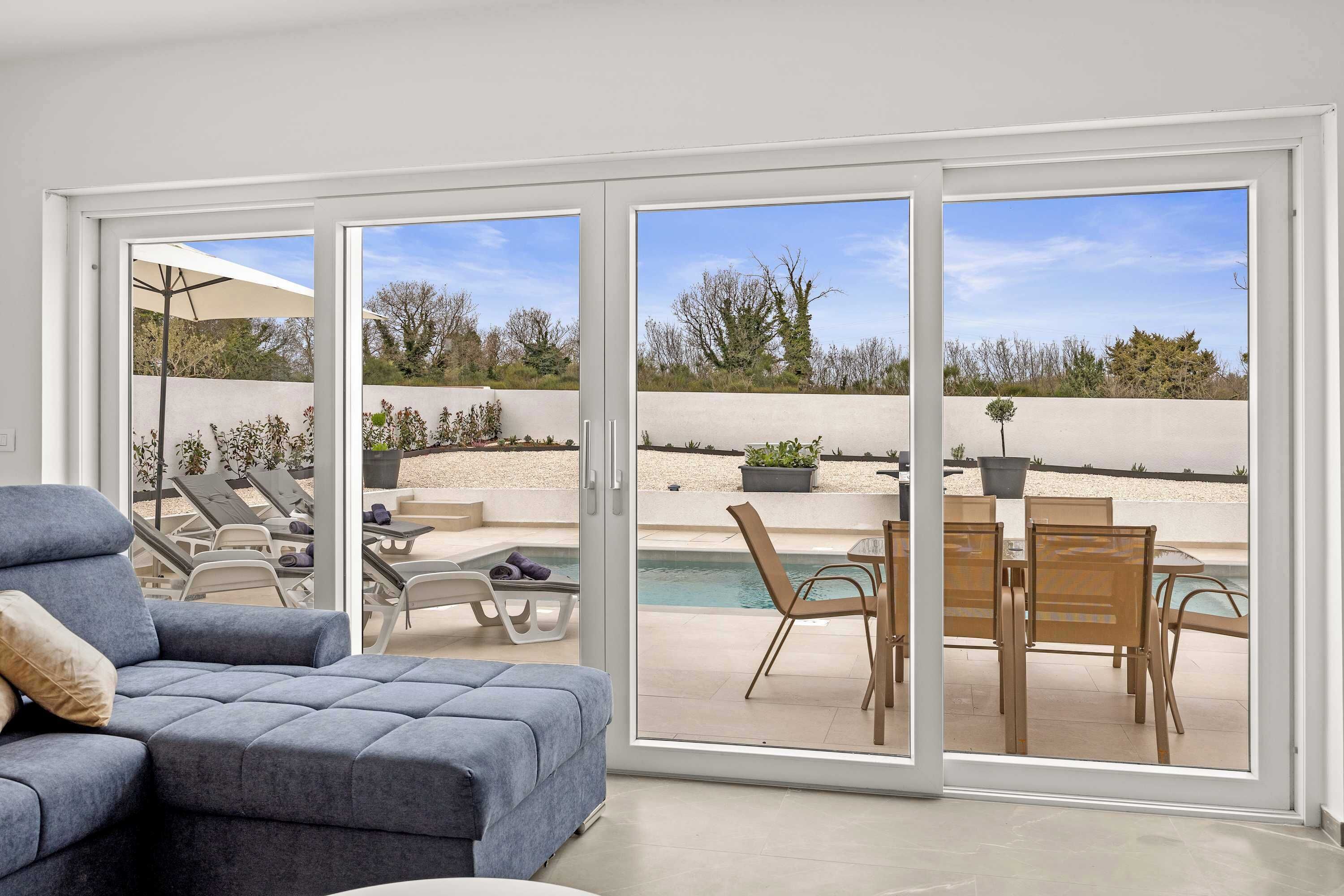 A modern living room with a grey sofa, a dining table with chairs, and a pool view through large sliding glass doors.