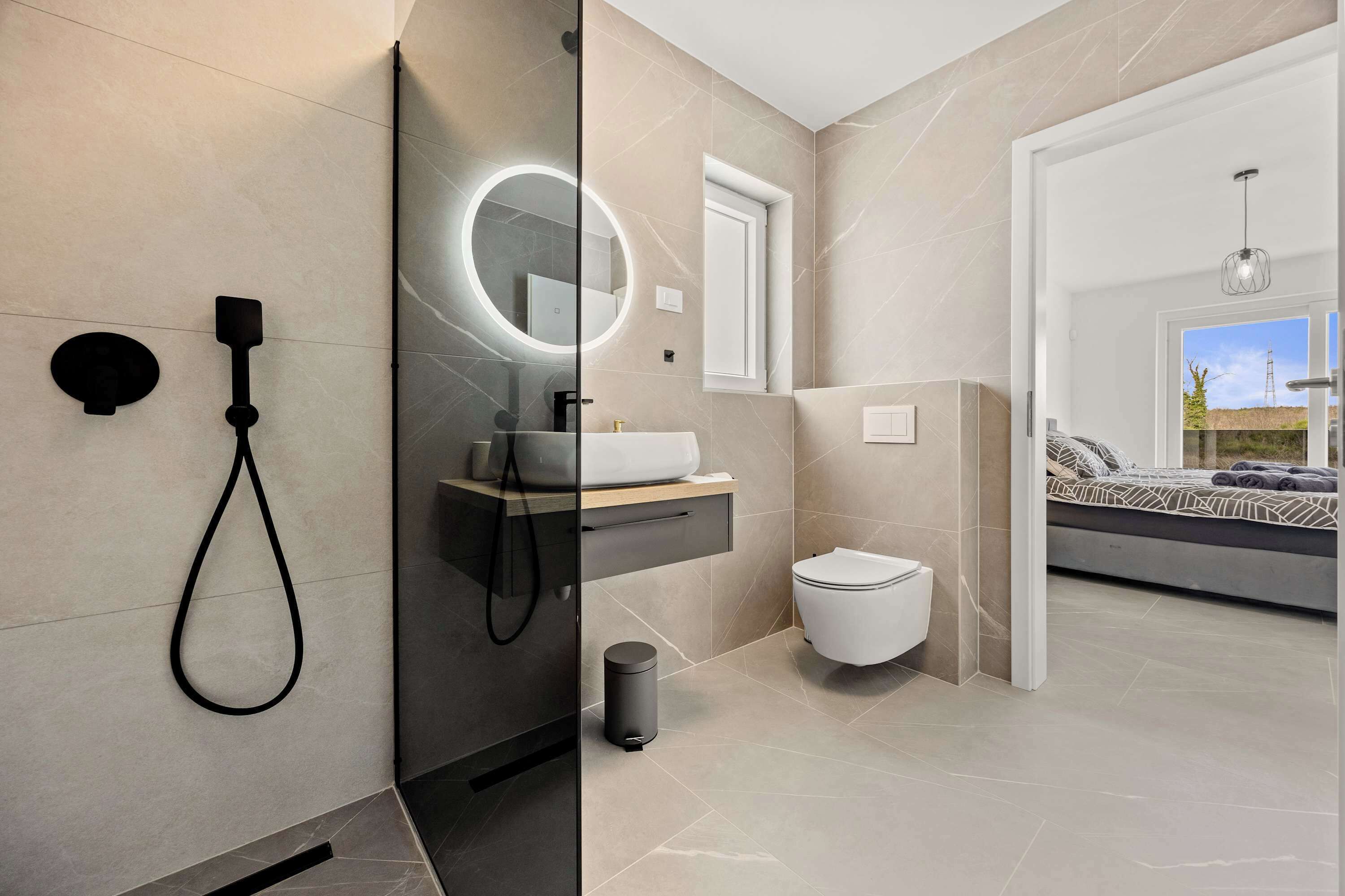 A modern bathroom with a glass-enclosed shower, a round mirror, a toilet, and a bed with a view.