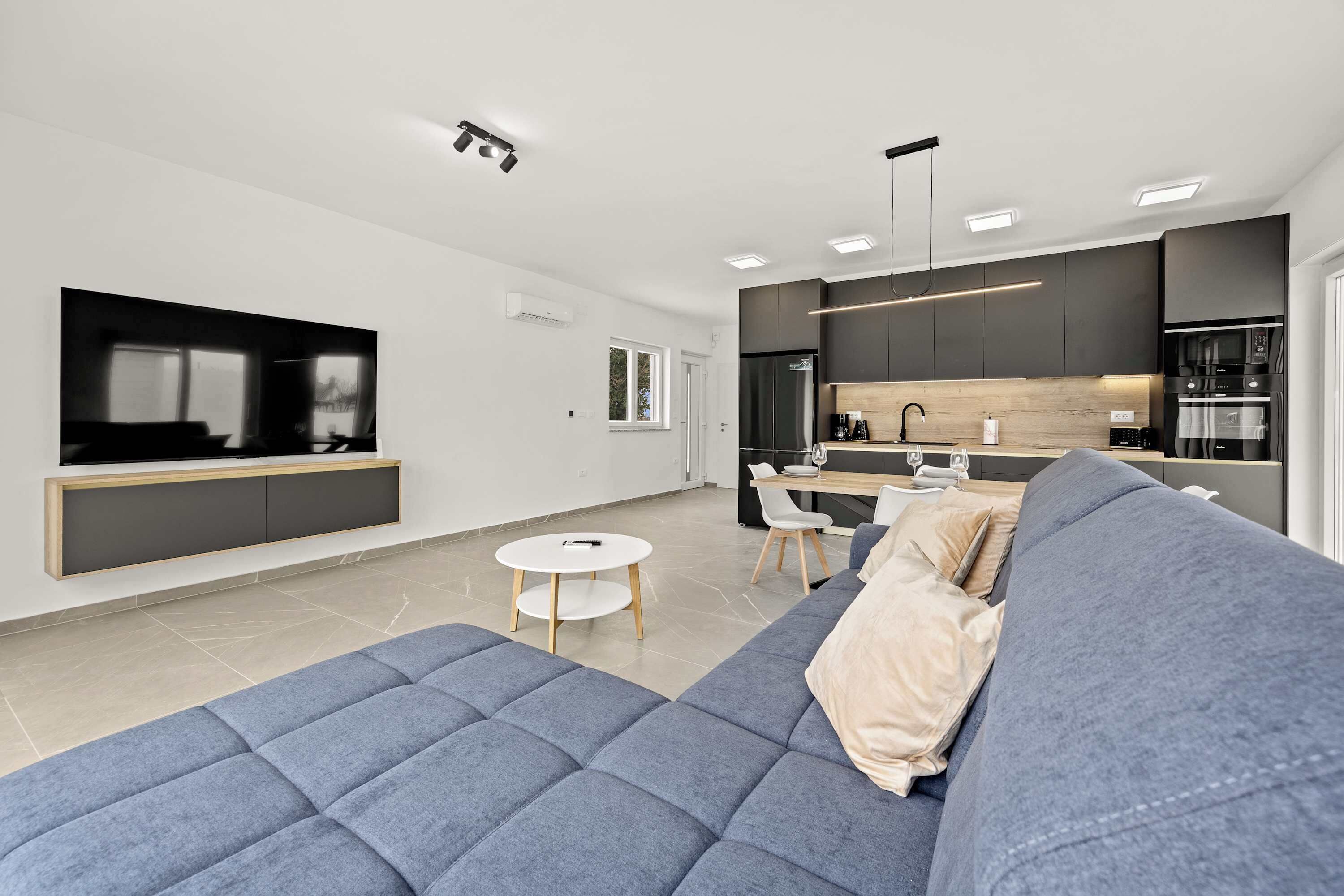 Een moderne woonkamer met een grote blauwe bank, een kleine ronde tafel en een keuken met donkere kasten en een houten aanrecht.