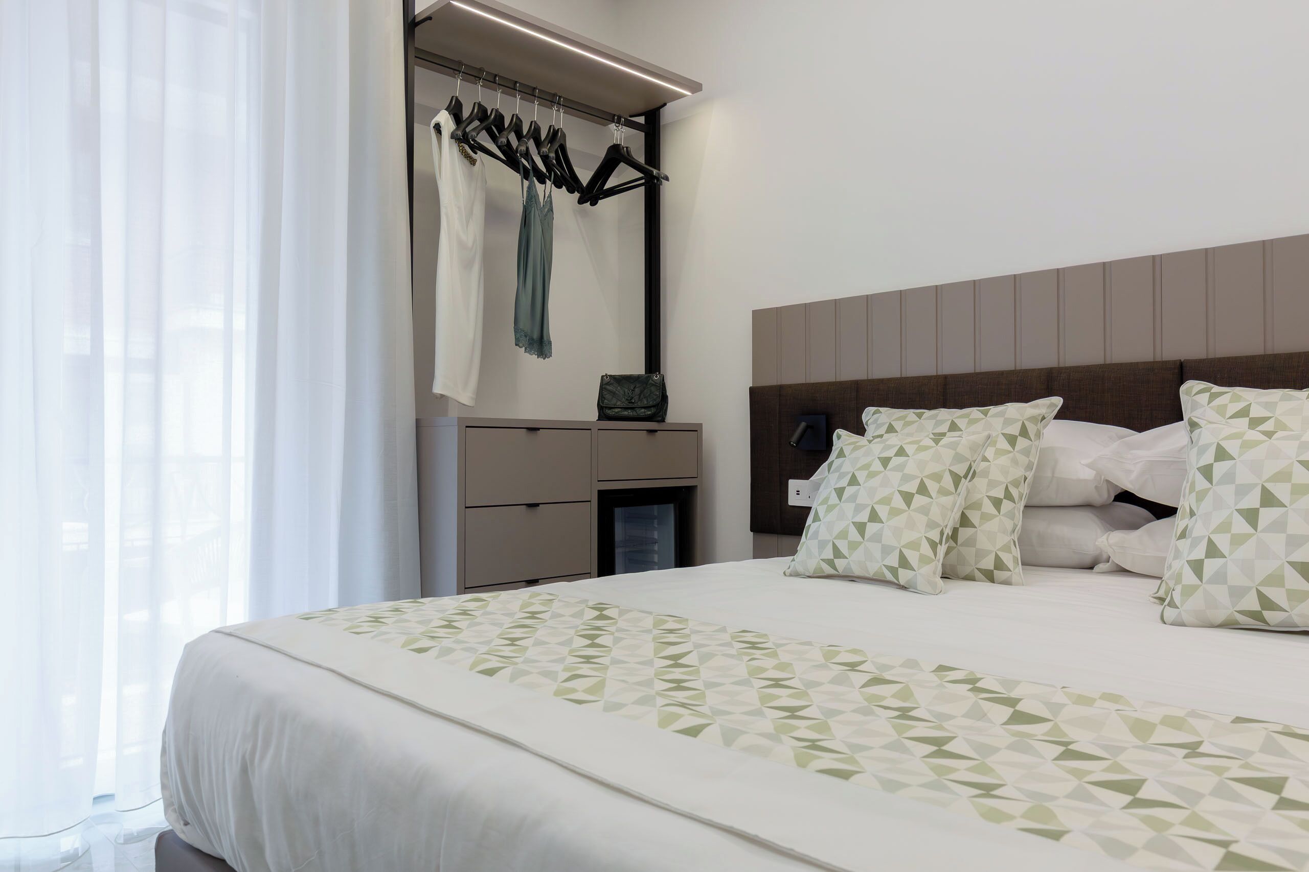Une chambre d’hôtel avec un lit, une armoire, un miroir et une fenêtre avec des rideaux.