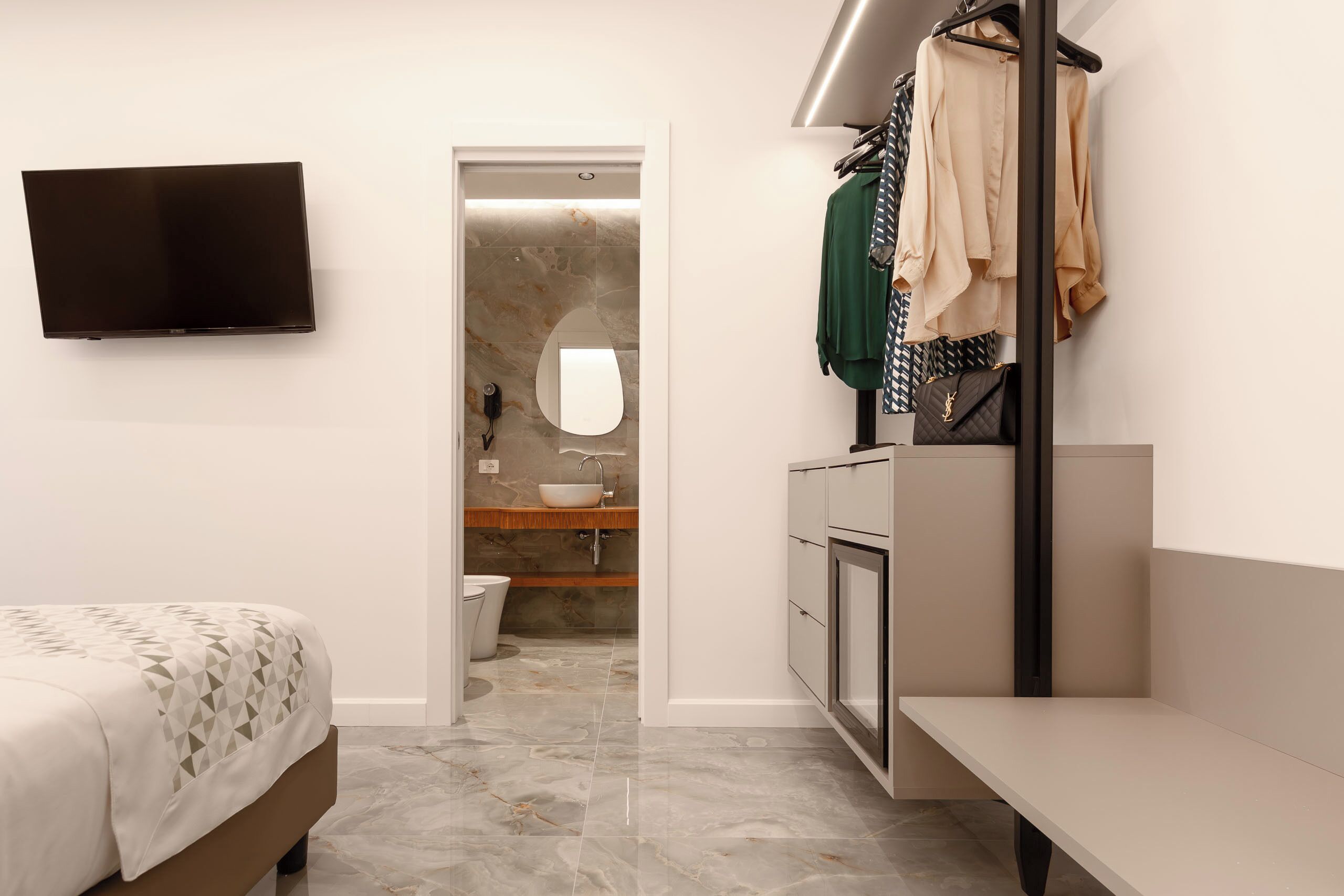 Une chambre moderne comprenant un lit, un téléviseur, une salle de bain avec un lavabo et un miroir, et un dressing avec des vêtements suspendus.