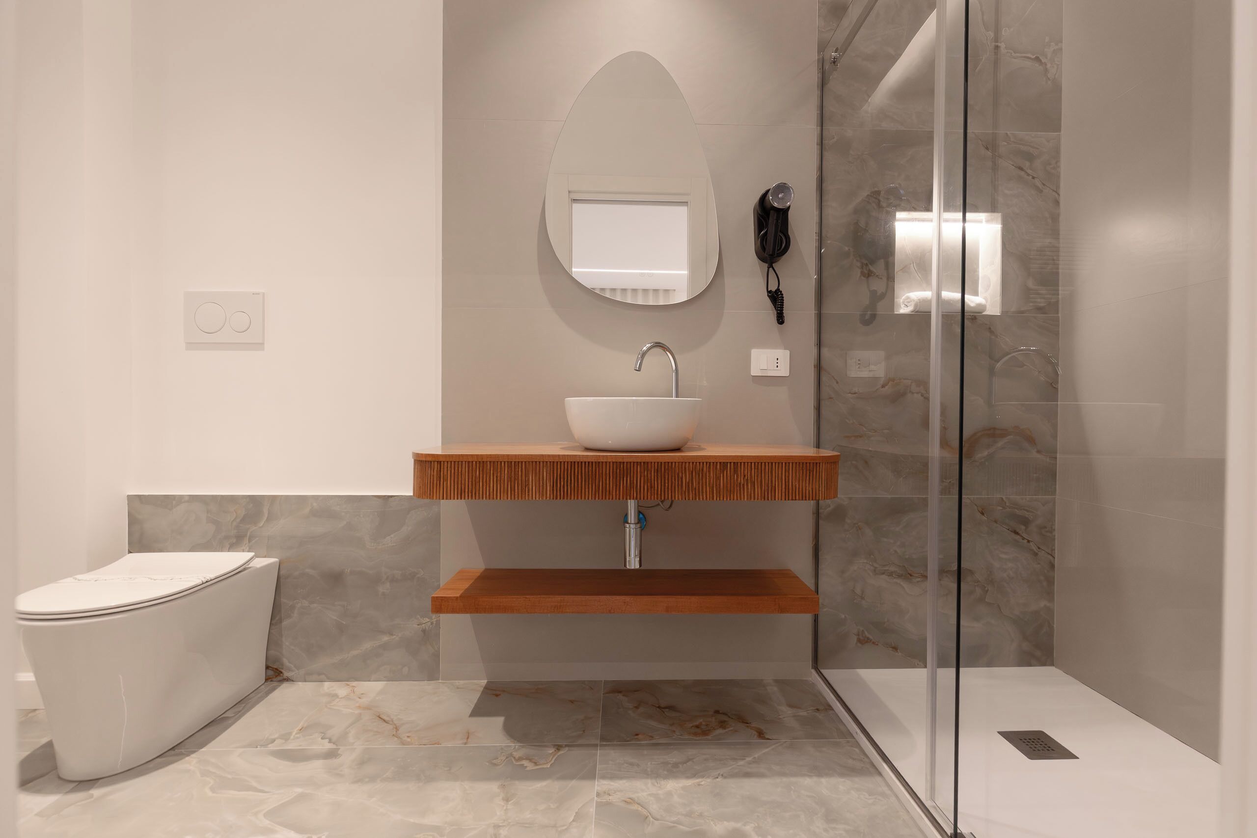A modern bathroom with a white toilet, a wooden vanity, a white sink, a mirror, a hairdryer, and a glass shower enclosure.