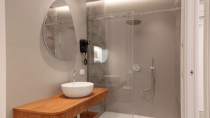 Un baño moderno con tocador de madera, un espejo redondo, un lavabo blanco y una ducha con mampara de vidrio.