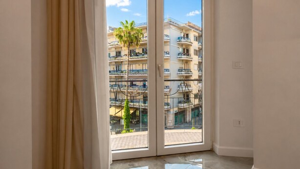Una habitación con un gran ventanal corredizo que da a un balcón con vistas a un edificio de varios pisos y una palmera.