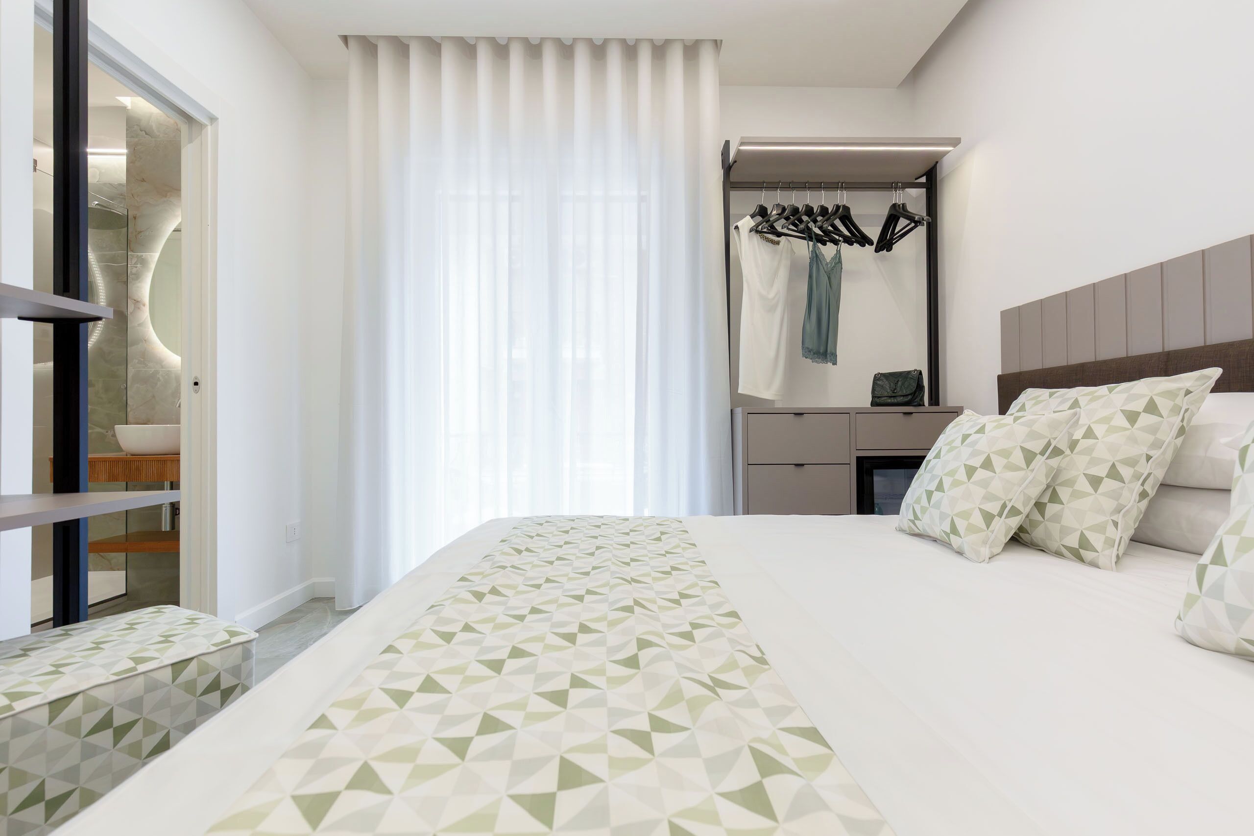 A modern hotel room with a large bed, a vanity with a mirror, and a clothing rack.