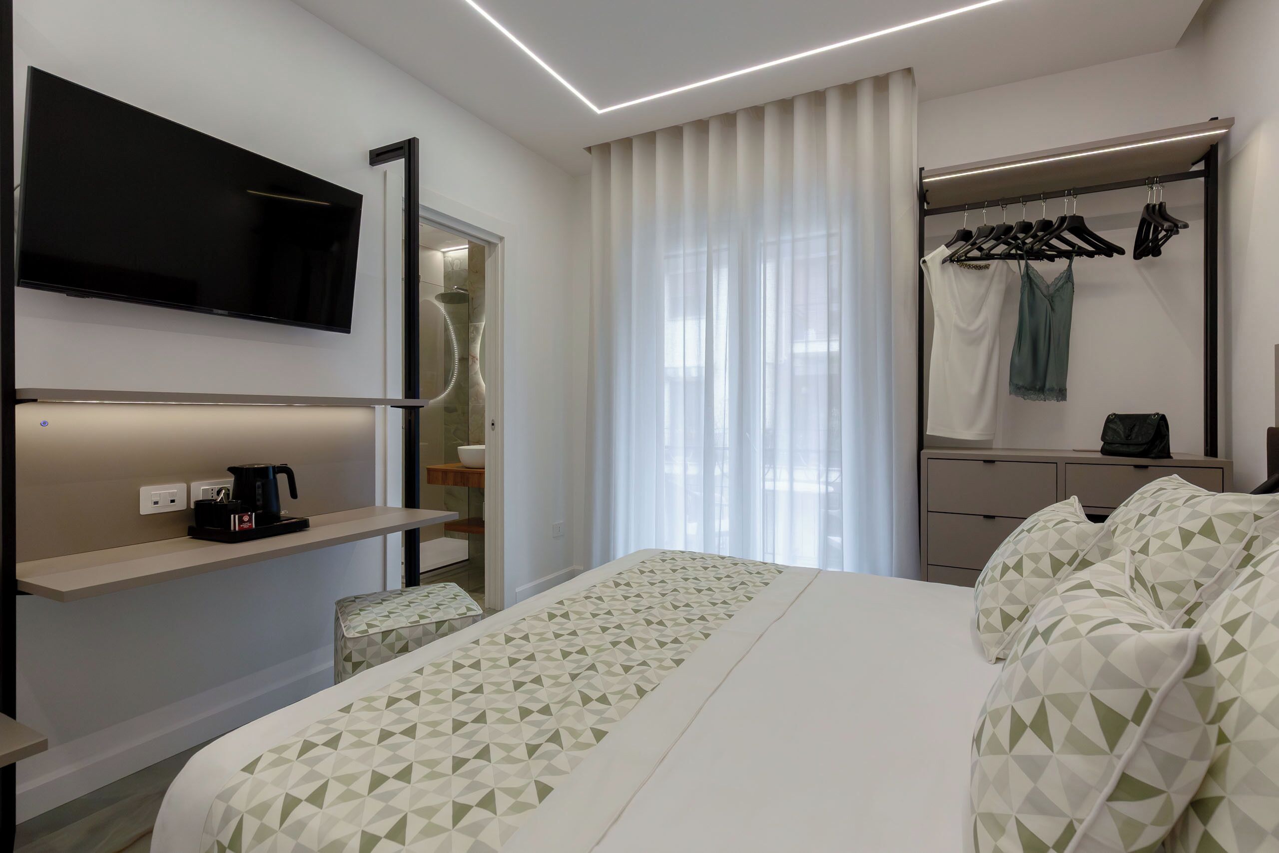 A hotel room with a bed, a TV mounted on the wall, a coffee maker, a wardrobe with hangers, and a window with curtains.
