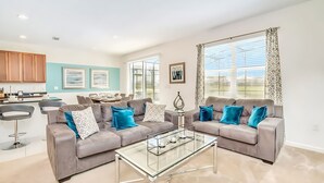 A modern living room with two grey sofas, a glass coffee table, and a kitchen in the background. - Upstay Lakefront Home w Pool Cinema Games BBQ (Kissimmee)