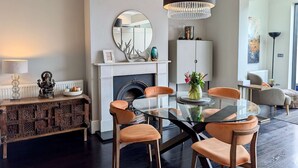 A modern living room with a fireplace, a round mirror, a chandelier, and a glass-top dining table with orange chairs. - 2 Private Ensuite Rooms Shared With Friendly Host (London)