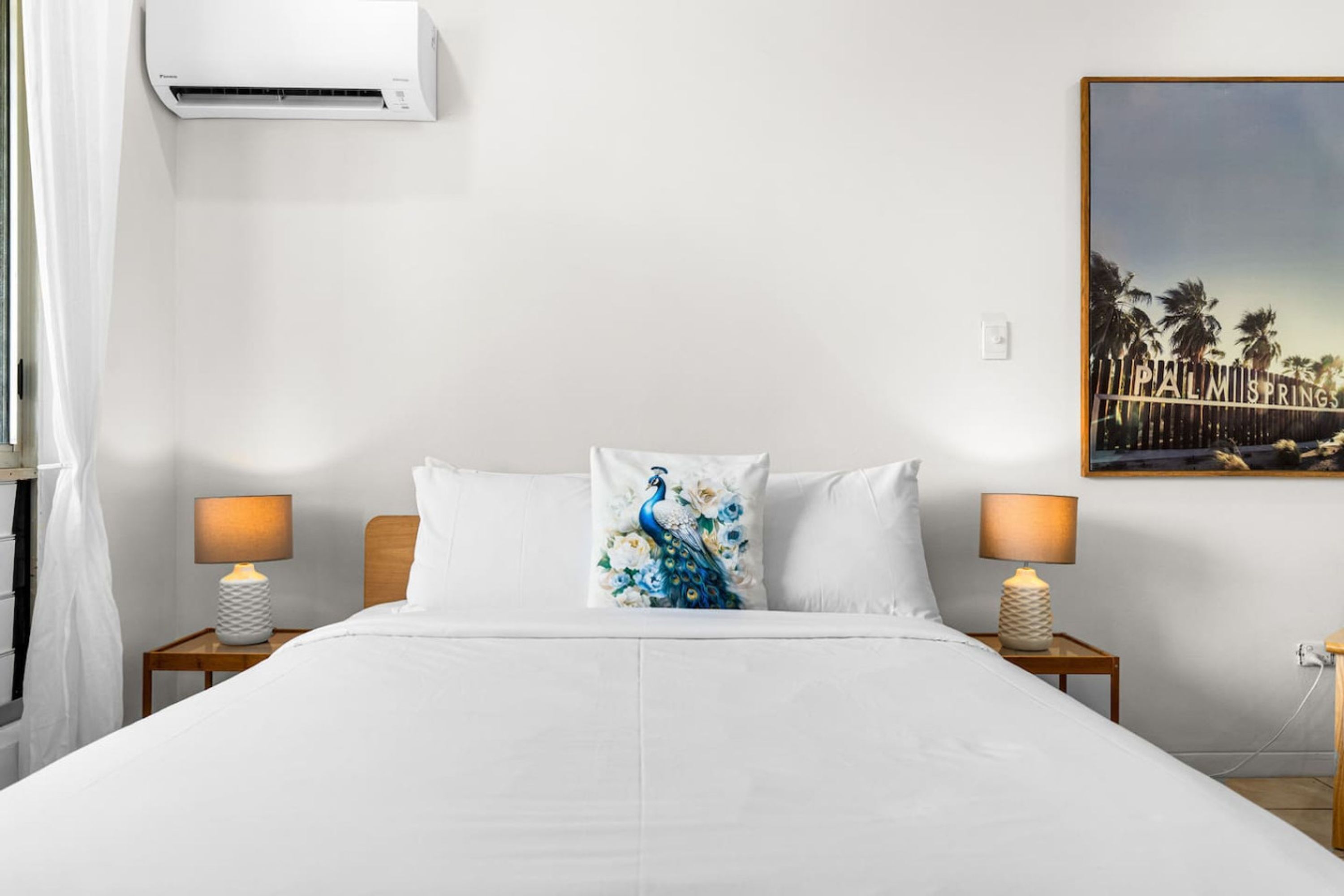 Ein Hotelzimmer mit einem Bett, zwei Nachttischen mit Lampen, einer Wand-Klimaanlage und einem gerahmten Bild an der Wand.