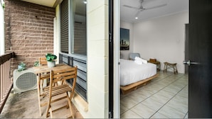 Une pièce comprenant un lit, une petite table, une chaise, une plante en pot et un balcon donnant sur un mur de briques.
